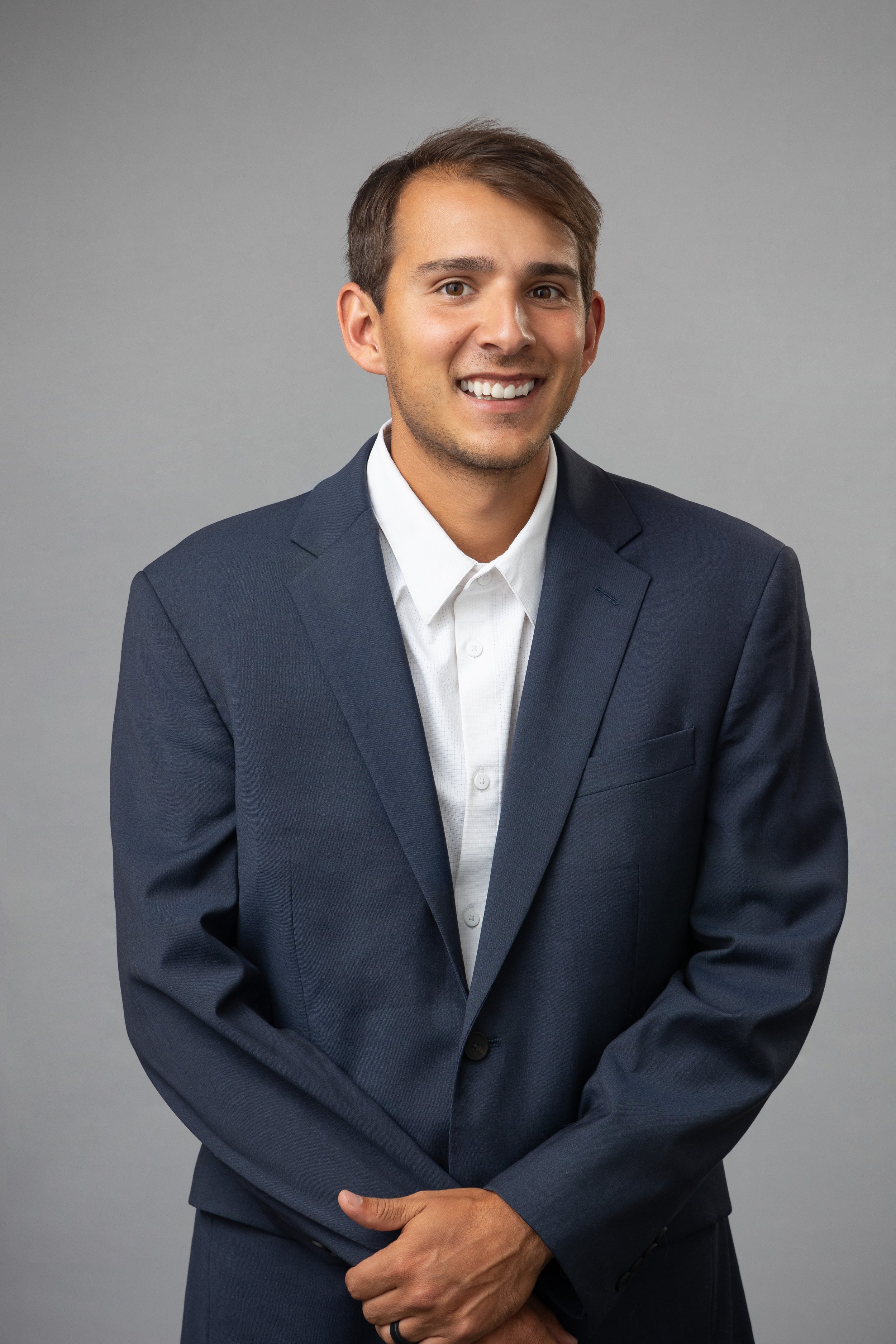 A young man in a navy suit and white shirt smiling at the camera against a plain gray background.