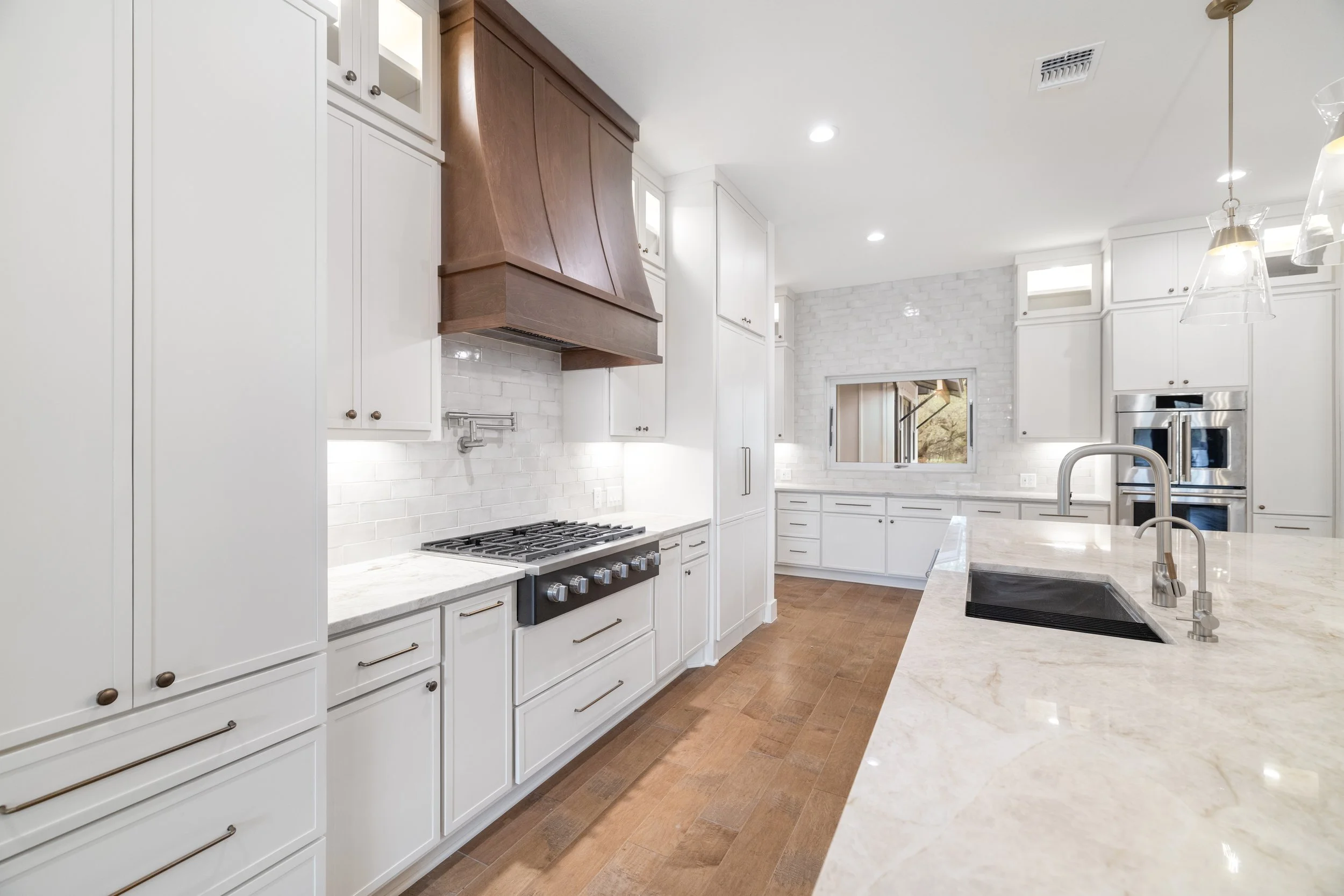 Modern white kitchen with marble countertops, wooden floors, and stainless steel appliances.