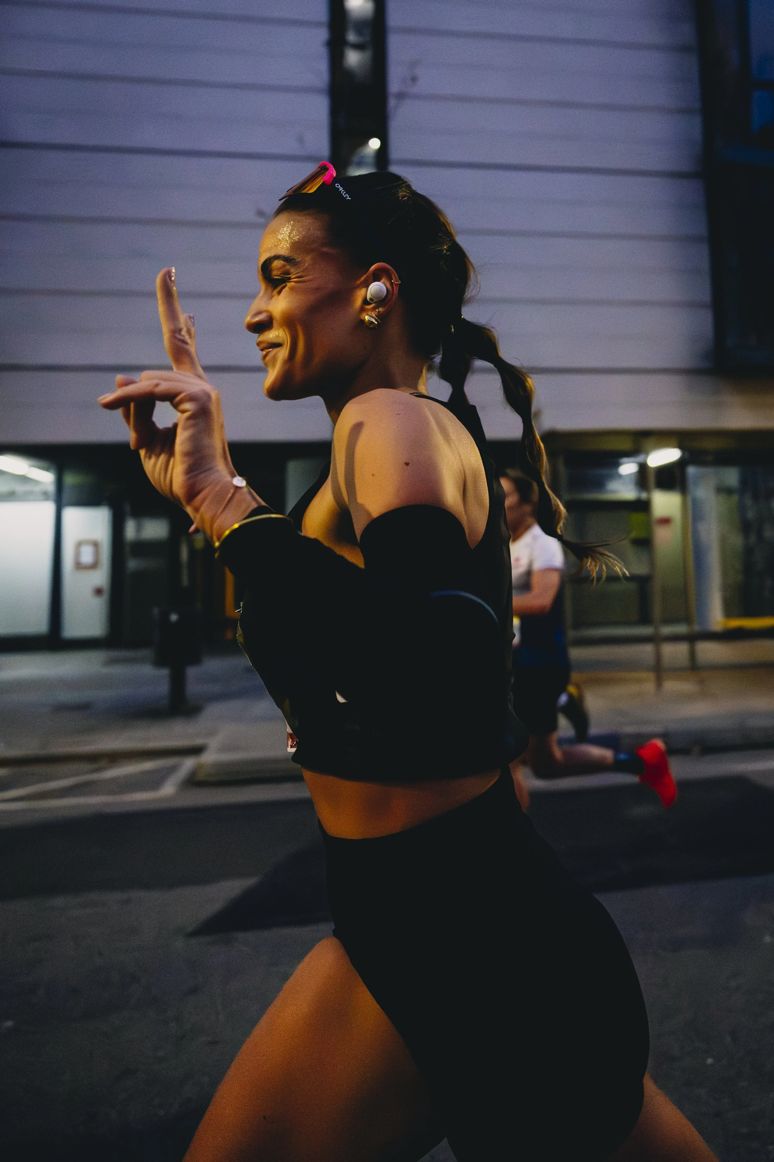 Mujer corriendo en la calle durante la noche, usando ropa deportiva negra, con auriculares blancos y gafas de sol en la cabeza.