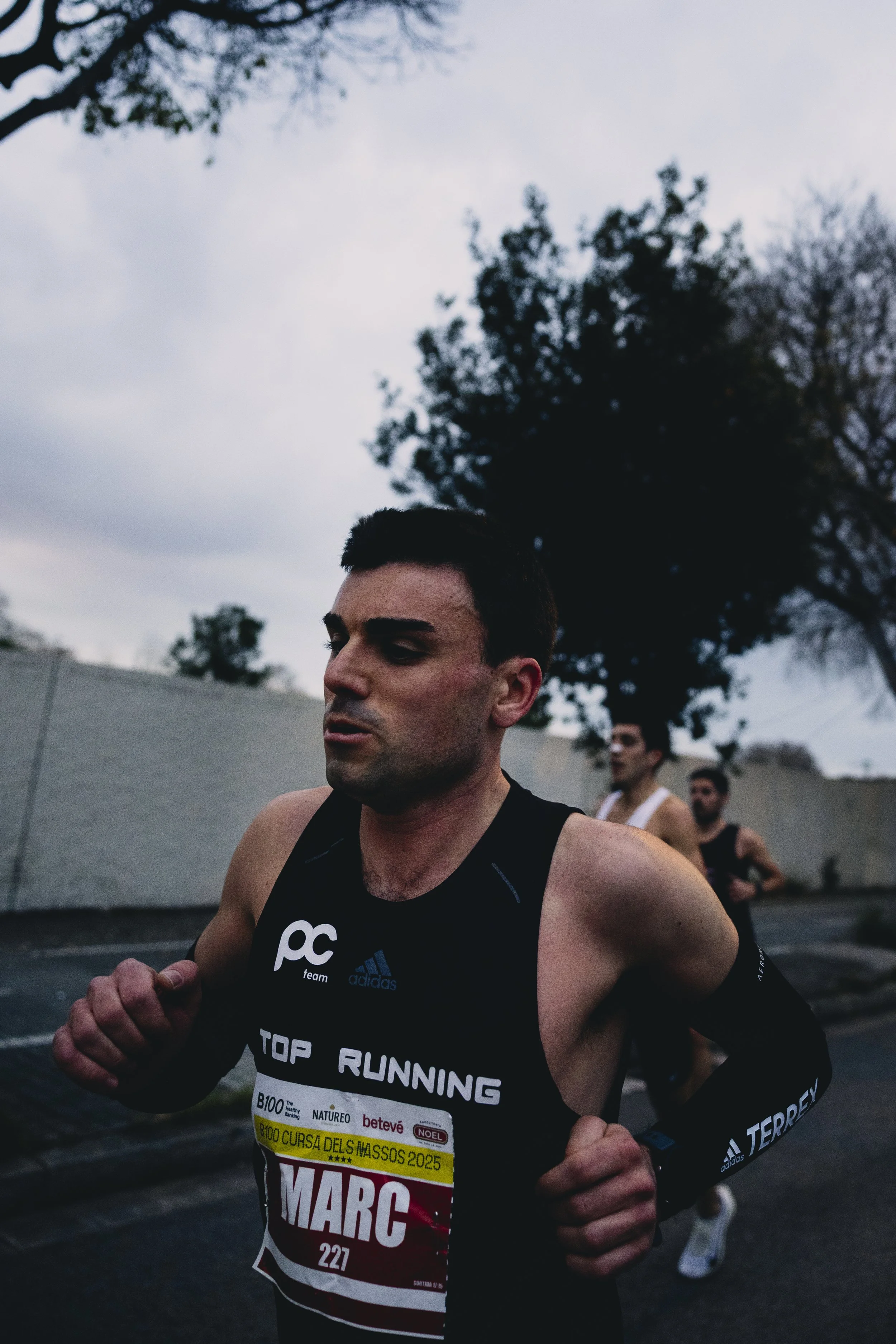 Hombre corriendo en una carrera de atletismo, llevando un uniforme negro con el número 227 y el nombre MARC, con otros corredores en el fondo y árboles en el cielo nublado.
