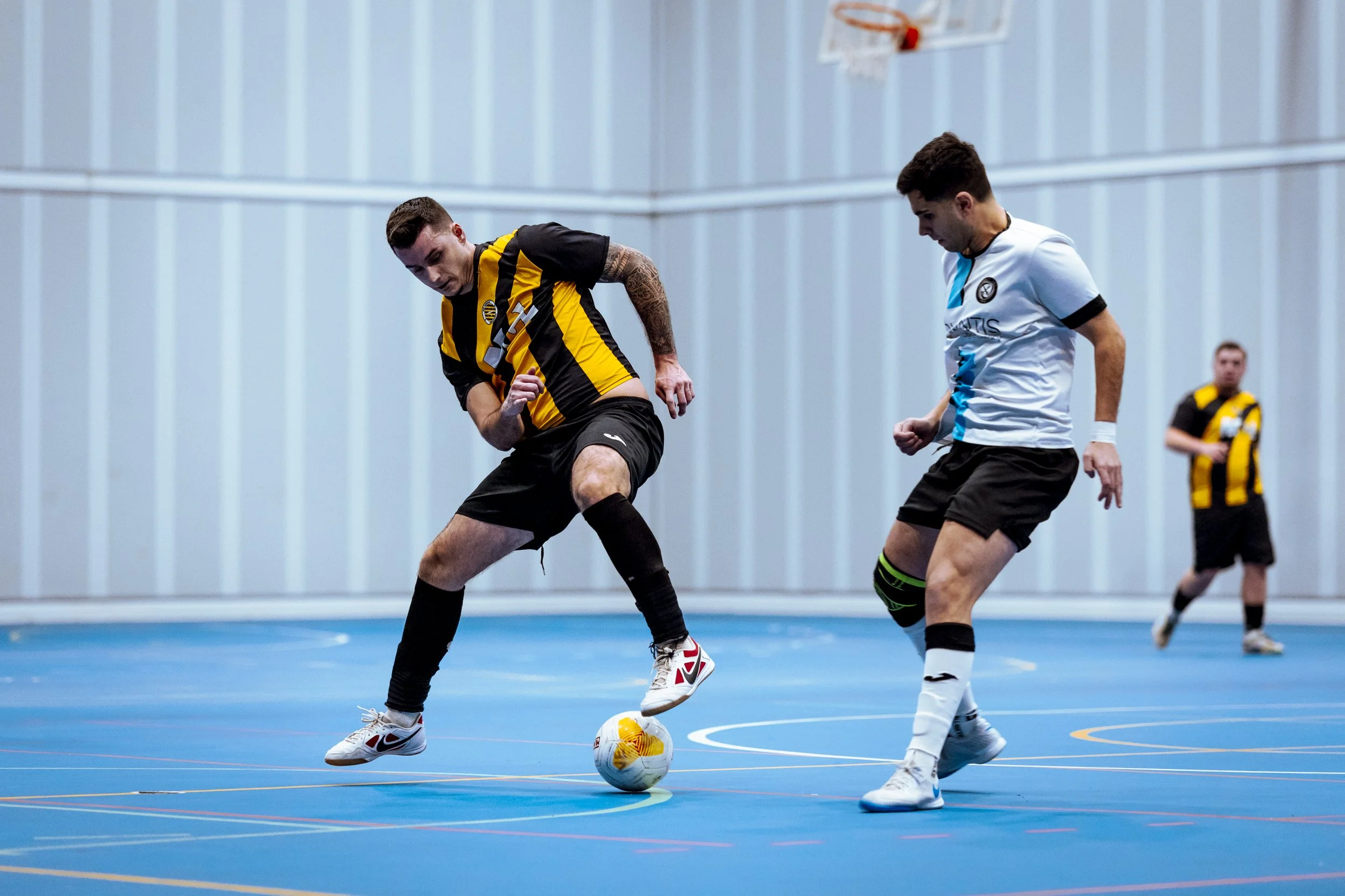 Dos jugadores de fútbol indoor compiten por el balón en una cancha azul, uno lleva uniforme amarillo y negro, y el otro lleva uniforme blanco y negro; hay un tercer jugador en el fondo.