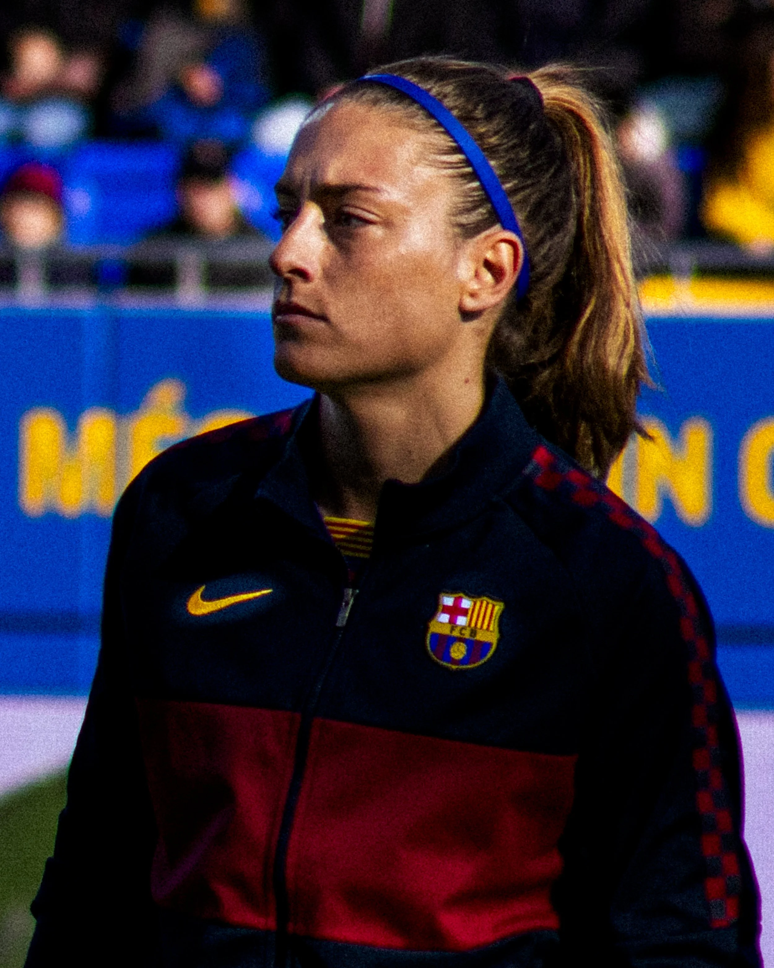 Una mujer con cabello en coleta atada con una diadema azul, lleva un uniforme delFC Barcelona, con la insignia y el logotipo, en un evento deportivo.