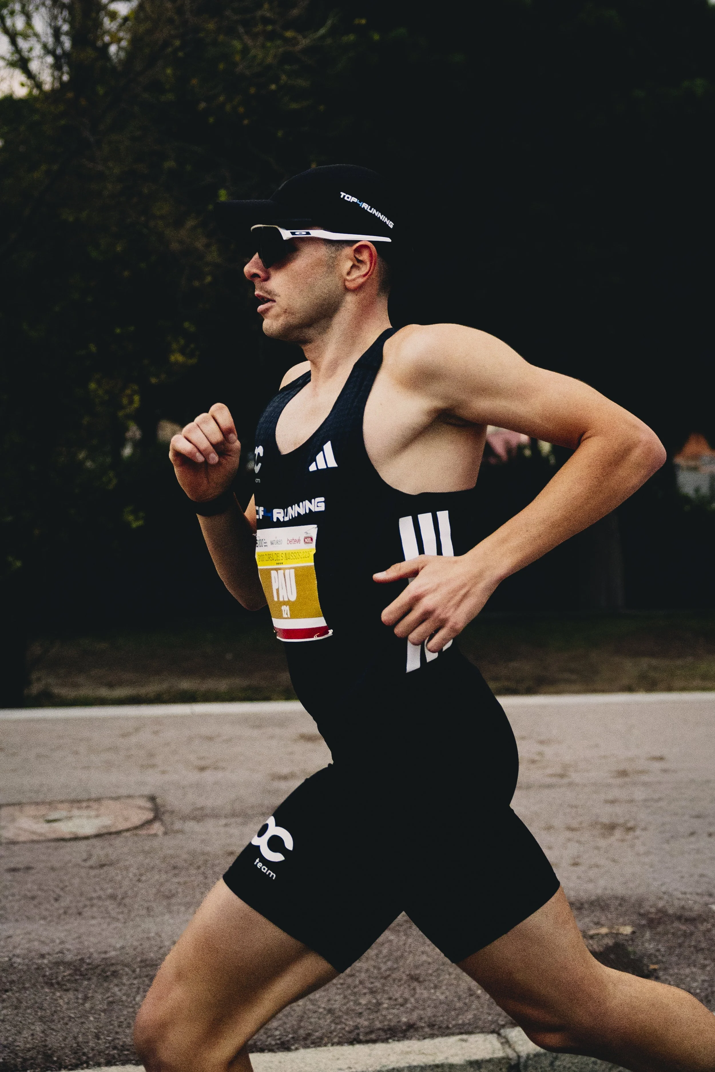 Hombre corriendo en una carrera de largas distancias, vestido con ropa deportiva negro, usando gafas de sol y gorra, en un fondo natural con árboles.