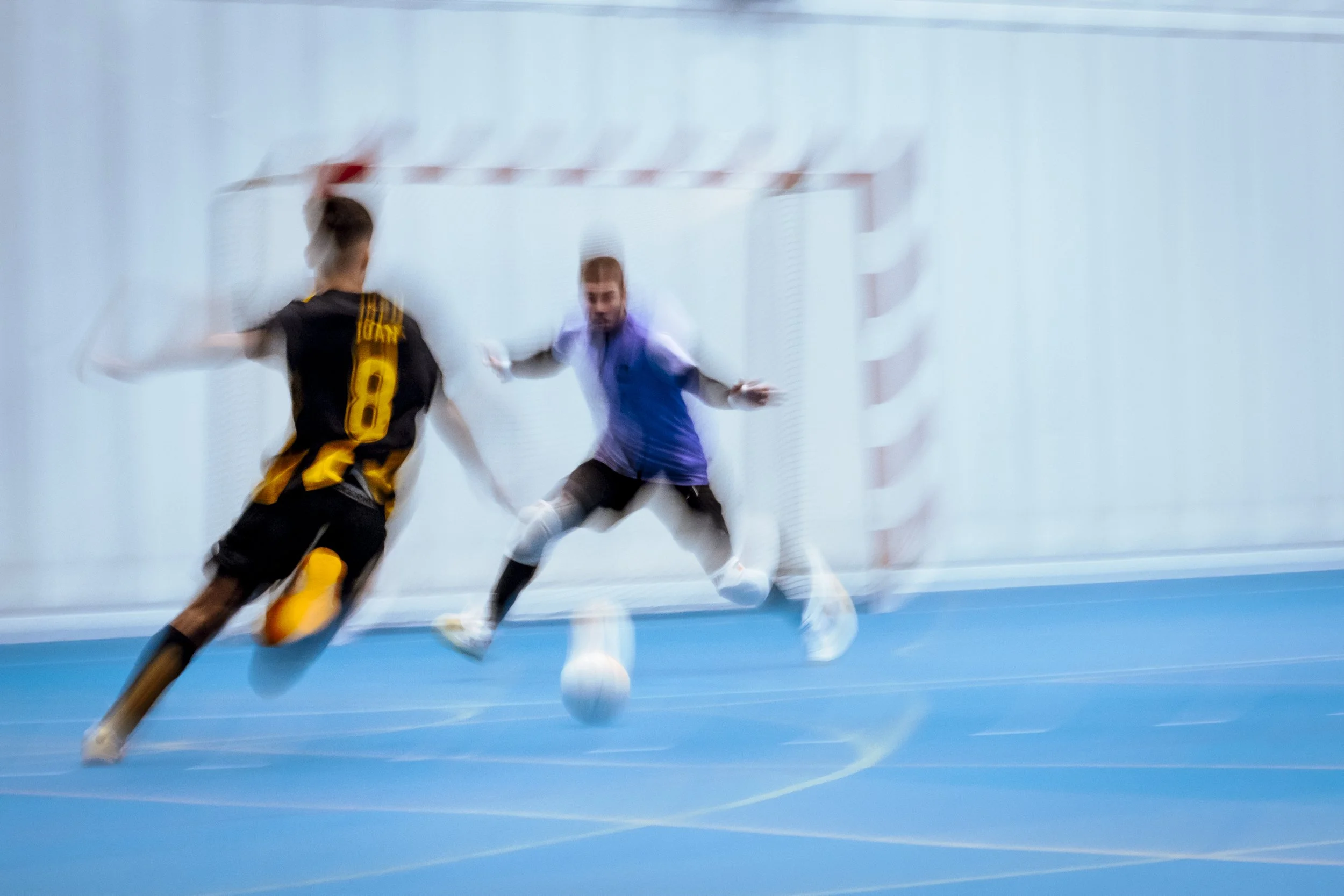 Dos jugadores de fútbol sala en acción, uno con camiseta negra y amarilla y otro con camiseta azul, en un intento de gol frente a la portería en una cancha cubierta.