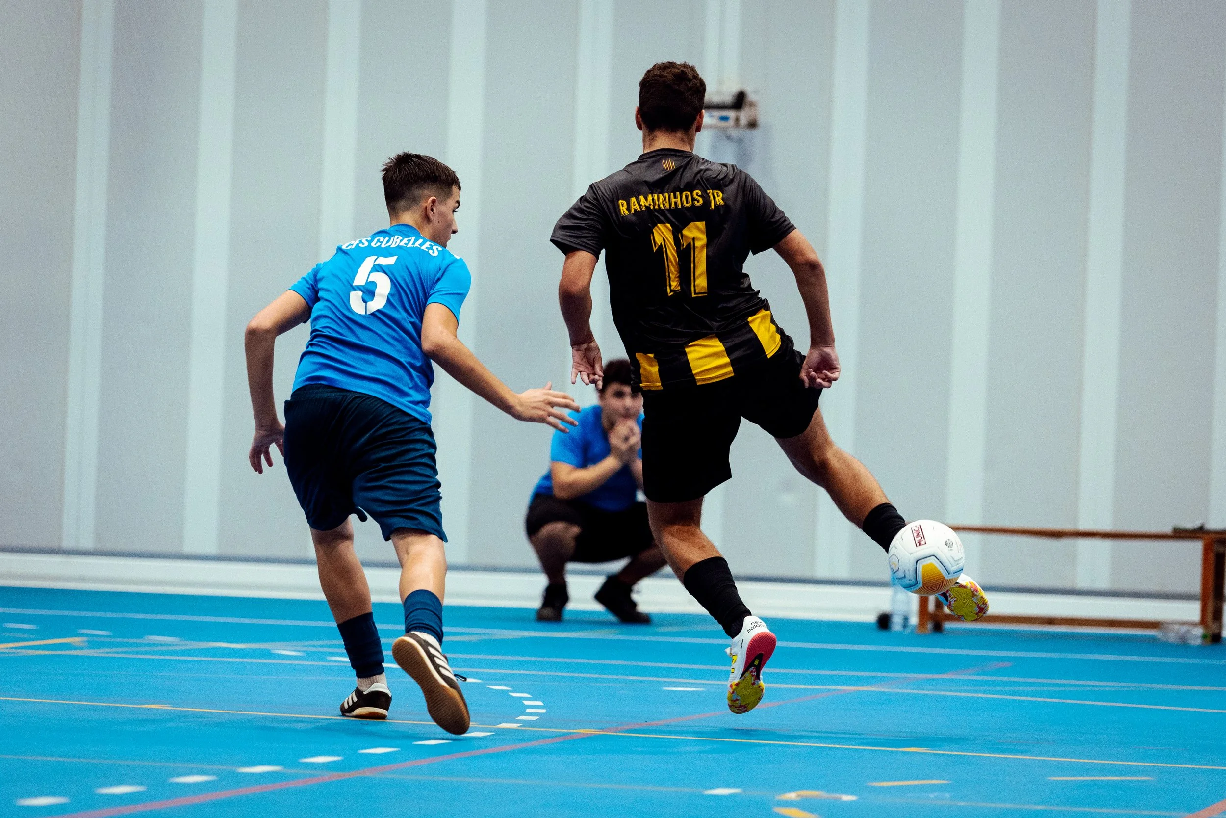Dos niños jugando futsal en una cancha cubierta, uno con uniforme azul y el otro con uniforme negro y amarillo, con un árbitro en el fondo observando.