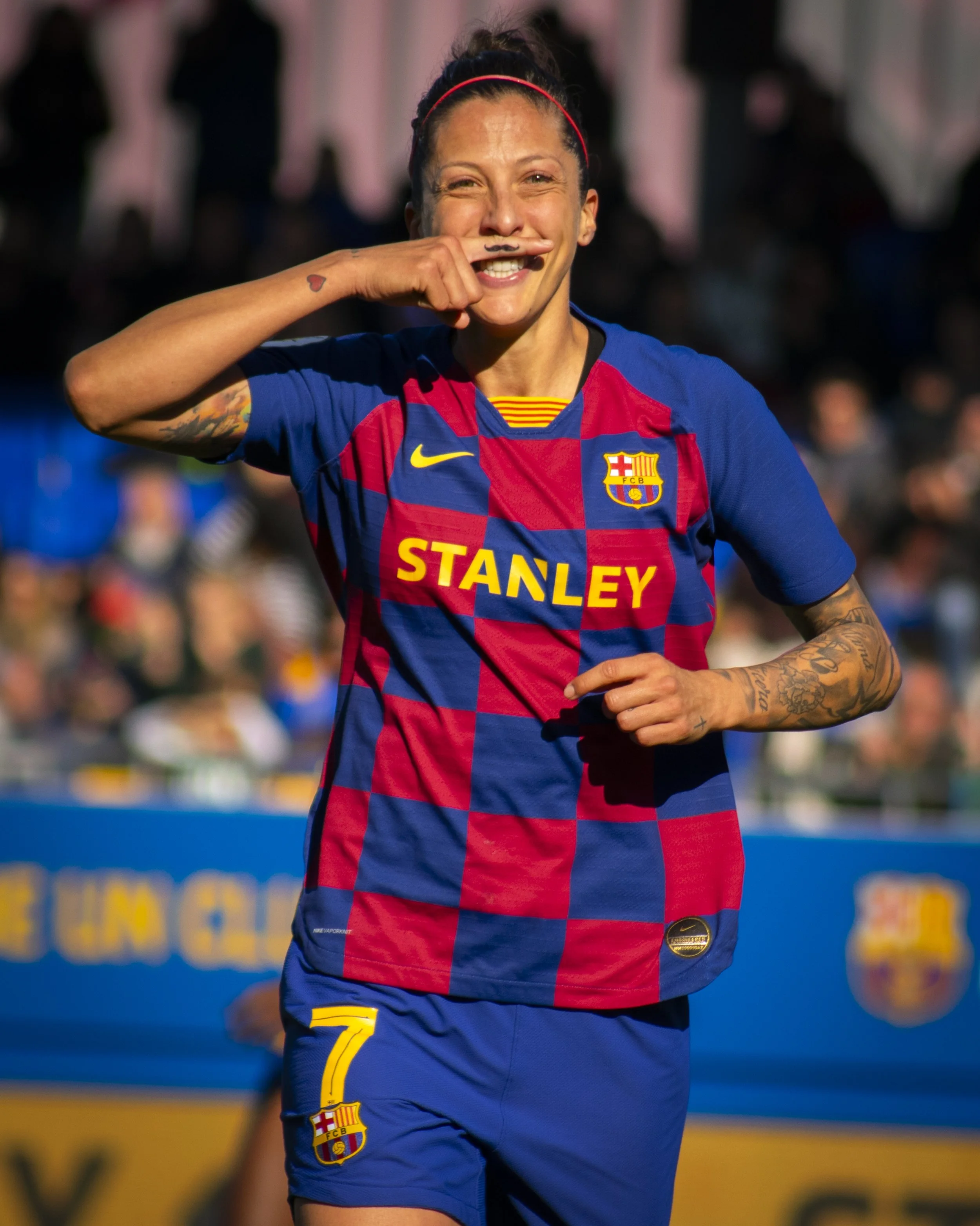 Una jugadora de fútbol en uniforma del FC Barcelona sonriendo y haciendo un gesto divertido con la cara, en un campo de fútbol, con público en el fondo.