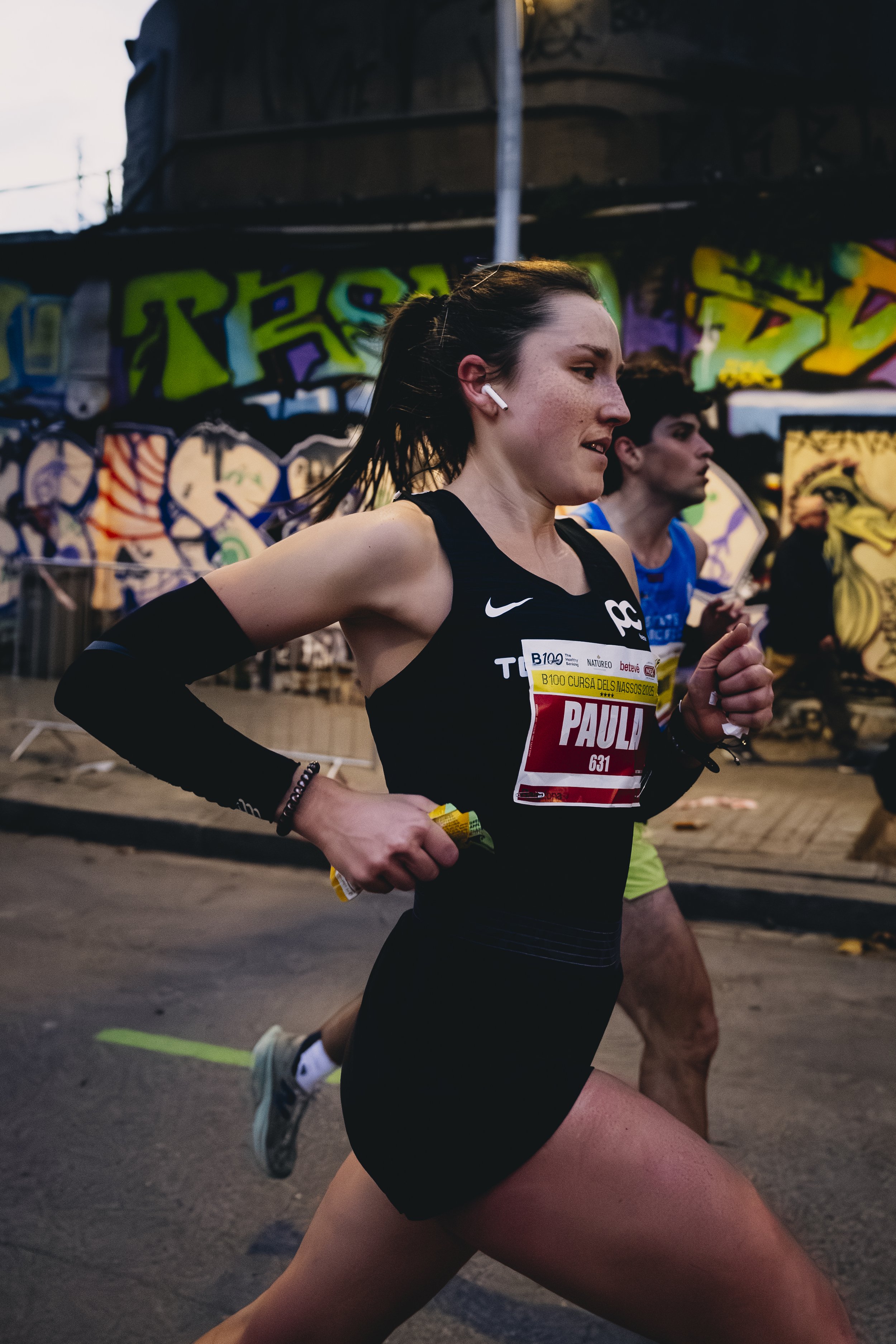 Mujer corriendo en una carrera, llevando audífonos, con un dorsal que dice 'PAULA' en un entorno urbano con graffiti de fondo.