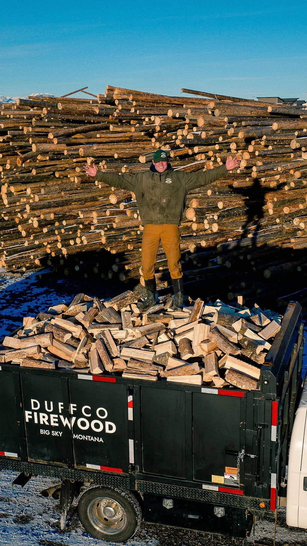 Duffco Firewood founder, Matt Duffy, standing on stacked firewood wearing green Duffco branded hat and jacket.