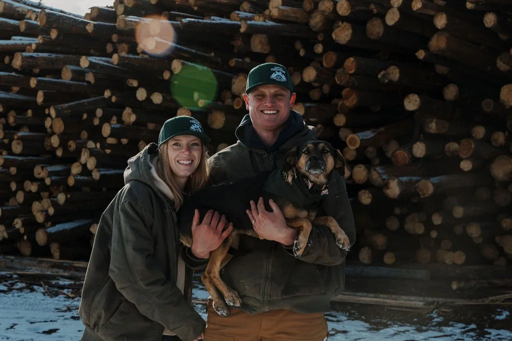 Duffco Firewood founders, Lyndsey and Matt Duffy, posing for a photo with stacked lumber behind them. Also, Duffco's Chief Morale Officer (Bella - dog) is in Matt's arms.
