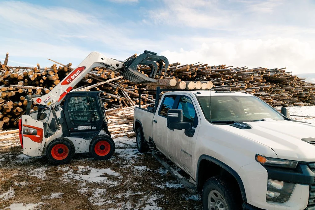 A bobcat loading posts & poles into the bed of a white truck.