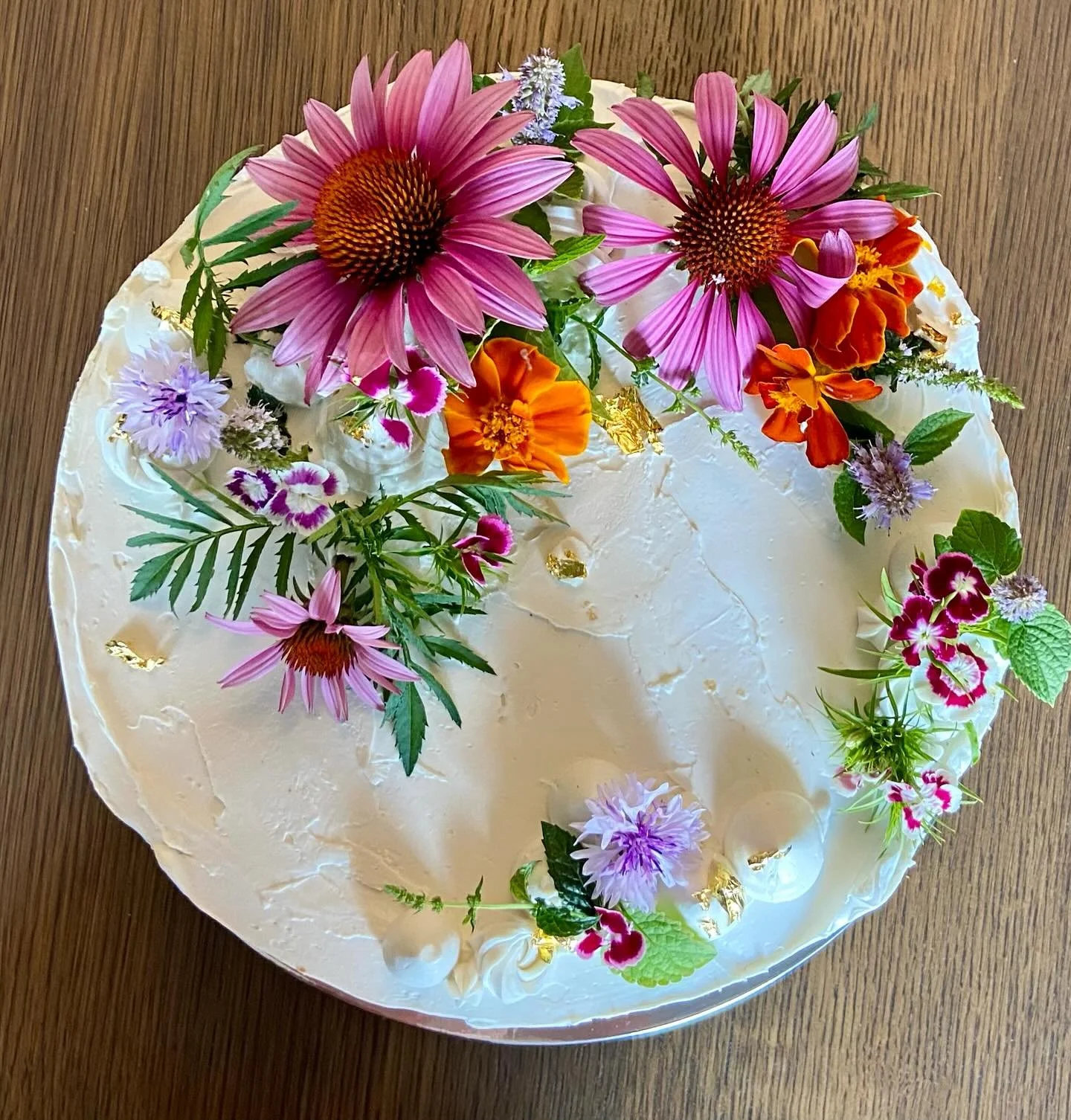 A white frosted cake decorated with colorful fresh flowers including purple and pink coneflowers, orange marigolds, and various small purple and pink garden flowers on a wooden surface.