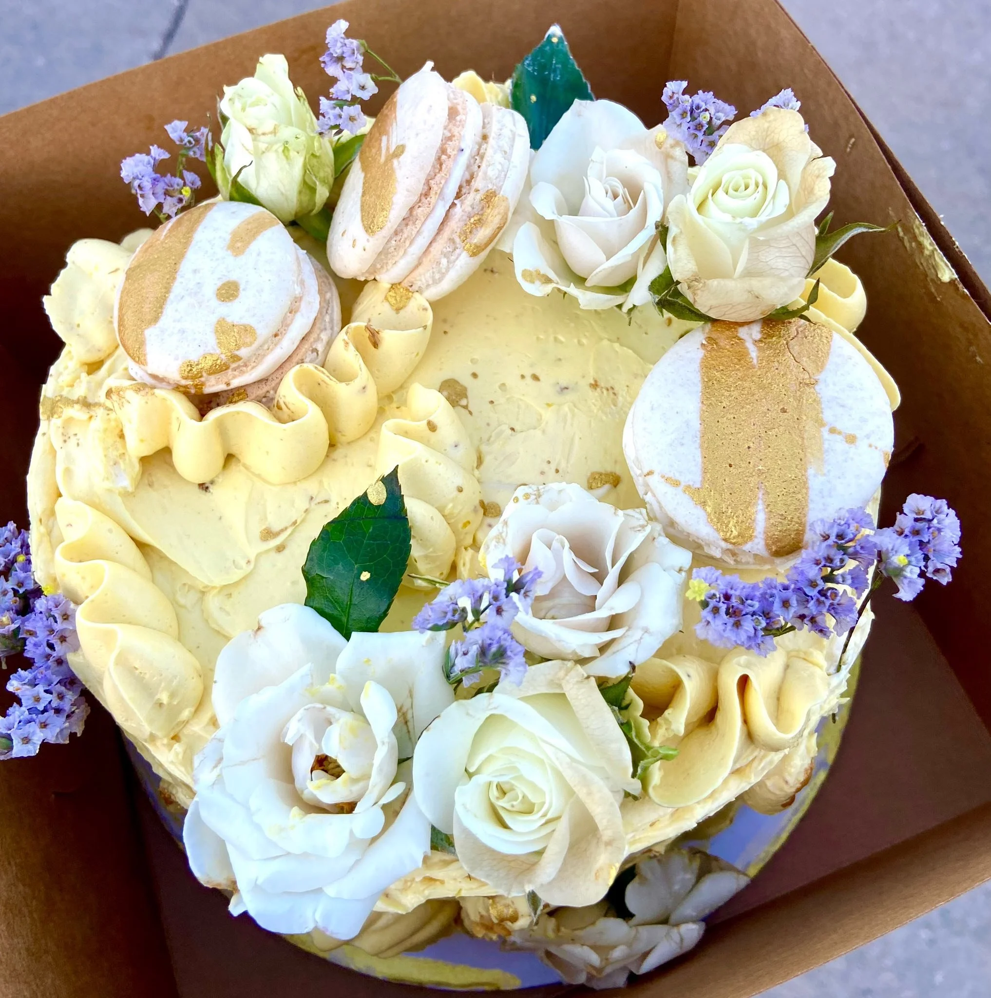 A decorative cake decorated with white roses, purple flowers, macarons, and yellow frosting.