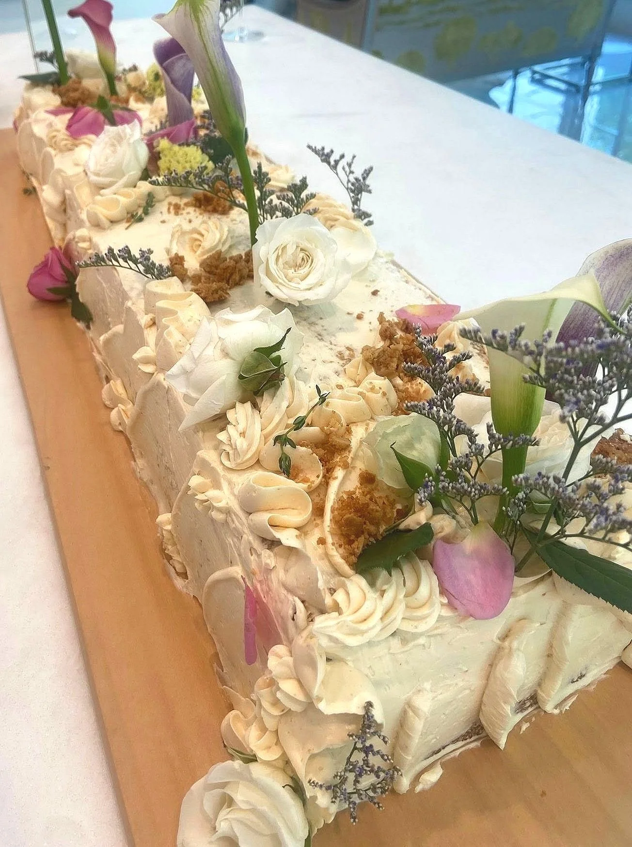 A rectangular wedding cake decorated with white and purple flowers, cream-colored frosting, and green leaves on a wooden board.