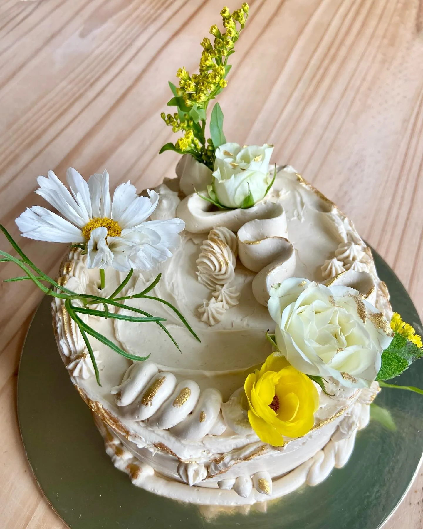 A decorated white cake with white, yellow, and green flowers on top, placed on a reflective silver cake board on a wooden surface.