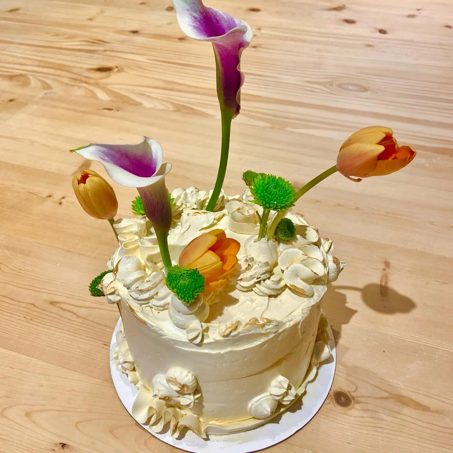 A round birthday cake decorated with white frosting, piped cream, and small cream rosettes around the edge, topped with artificial flowers including purple calla lilies, orange tulips, and small green decorative elements, all on a wooden surface.