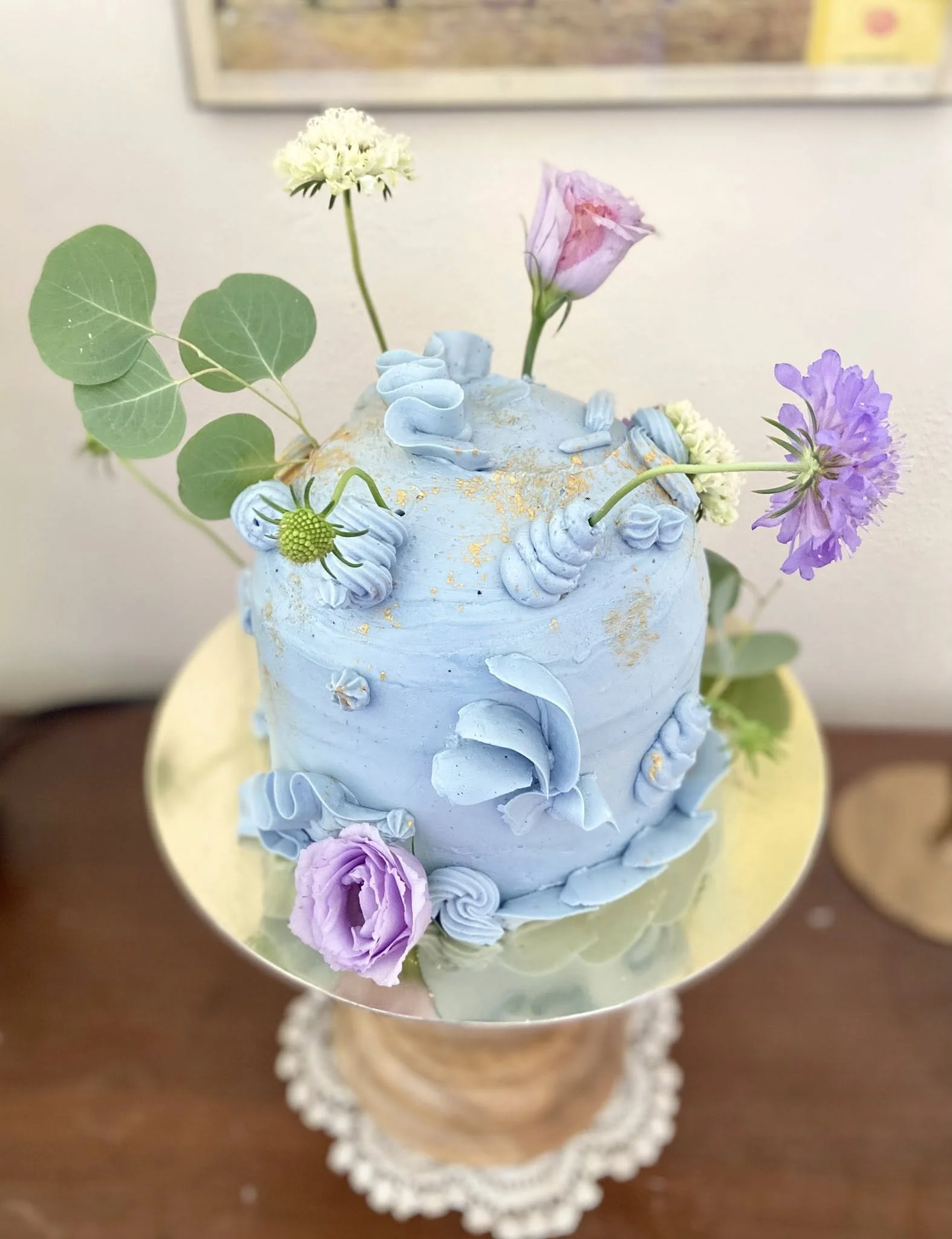A light blue frosted cake decorated with fresh flowers, including purple and pink roses, and eucalyptus leaves. The cake is on a gold cake board and placed on a wooden surface with a decorative doily underneath.