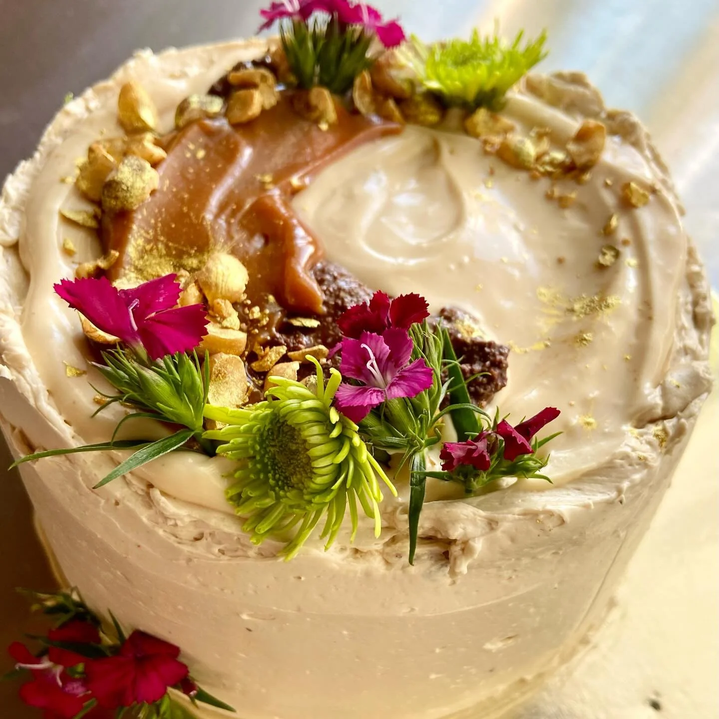 A decorated cake with white frosting, edible flowers, nuts, gold leaf, caramel, and chocolate toppings.