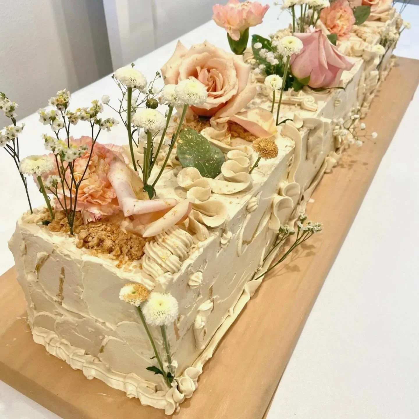 A rectangular cake decorated with pink and white flowers, green leaves, white frosting, and various edible and decorative toppings on a wooden board.