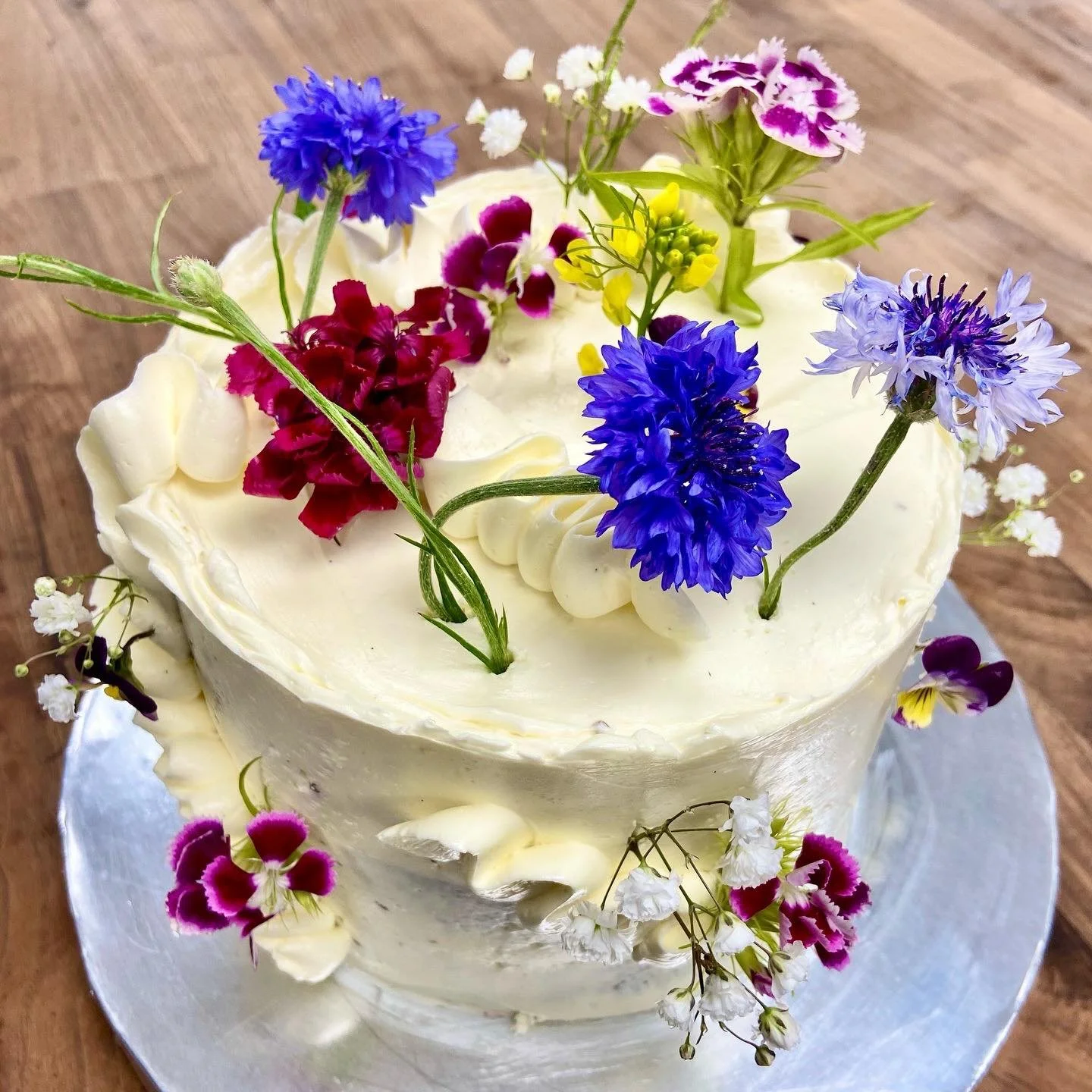 A white cake decorated with fresh mixed flowers including cornflowers and small white and purple blooms, on a wooden surface.