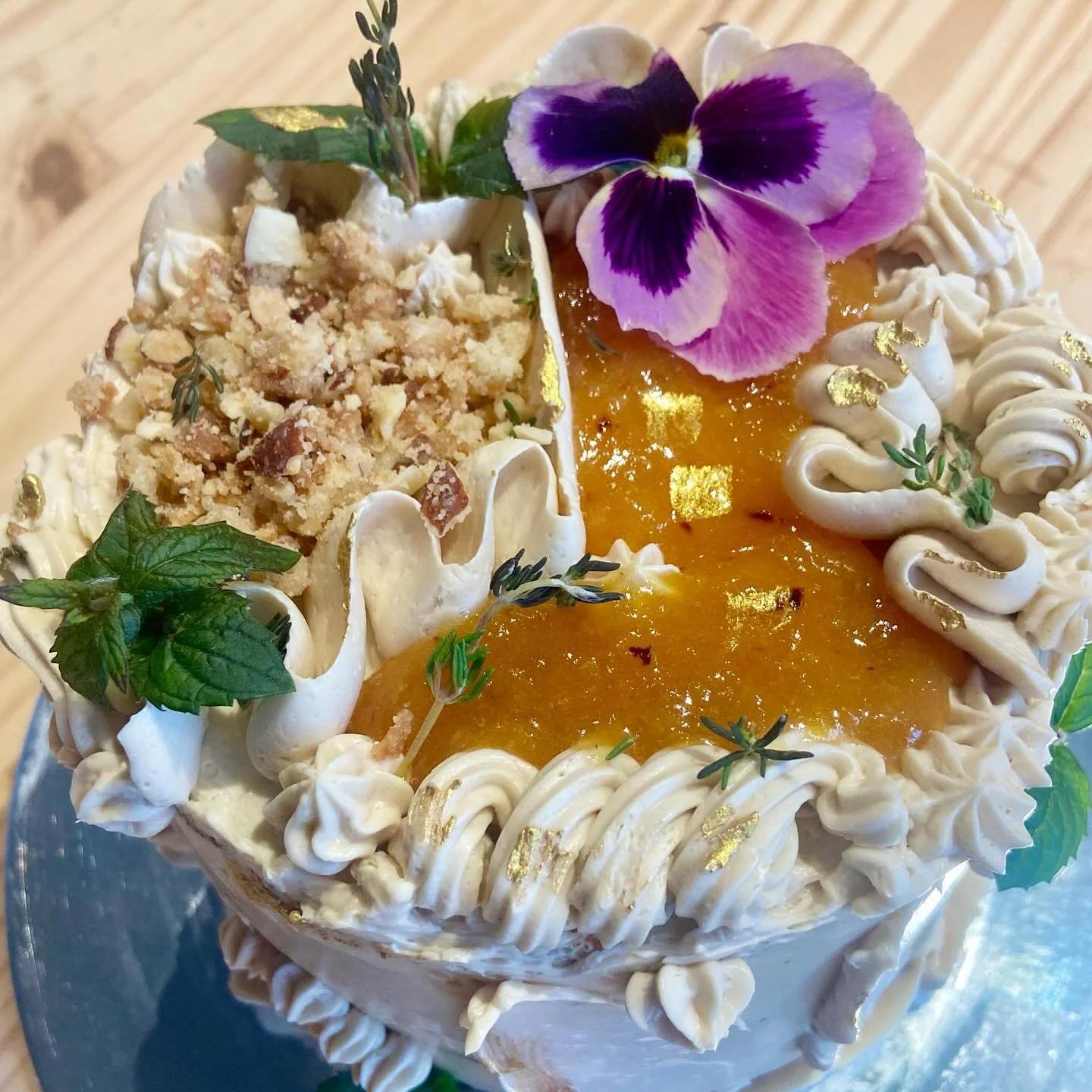 Decorated cake with white frosting, edible gold leaf, purple and yellow edible flowers, and chopped nuts, garnished with sprigs of herbs.