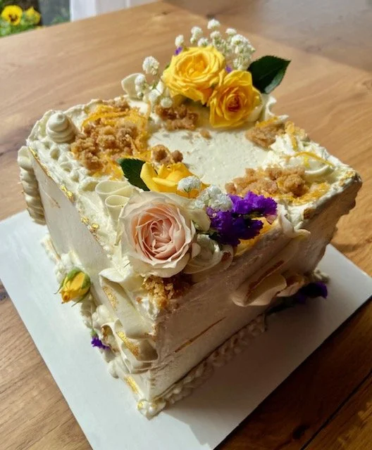 Square cream-colored cake decorated with yellow and pink roses, purple flowers, and white baby's breath, topped with crumbs and floral accents, on a white base on a wooden table.
