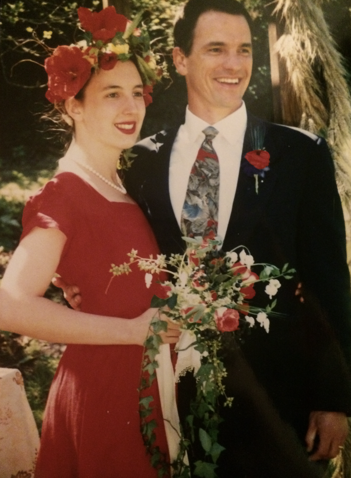 Ken and Ellen on their wedding day, smiling together outdoors; Ellen wears a red dress with a floral headpiece, reflecting her background as a florist.