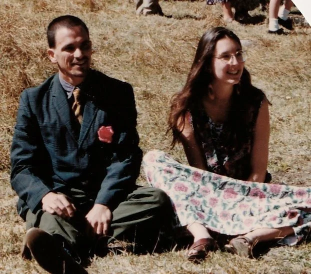 Ken and Ellen, founders of MAK, sitting together outdoors on dry grass, smiling in casual vintage attire before starting their business.