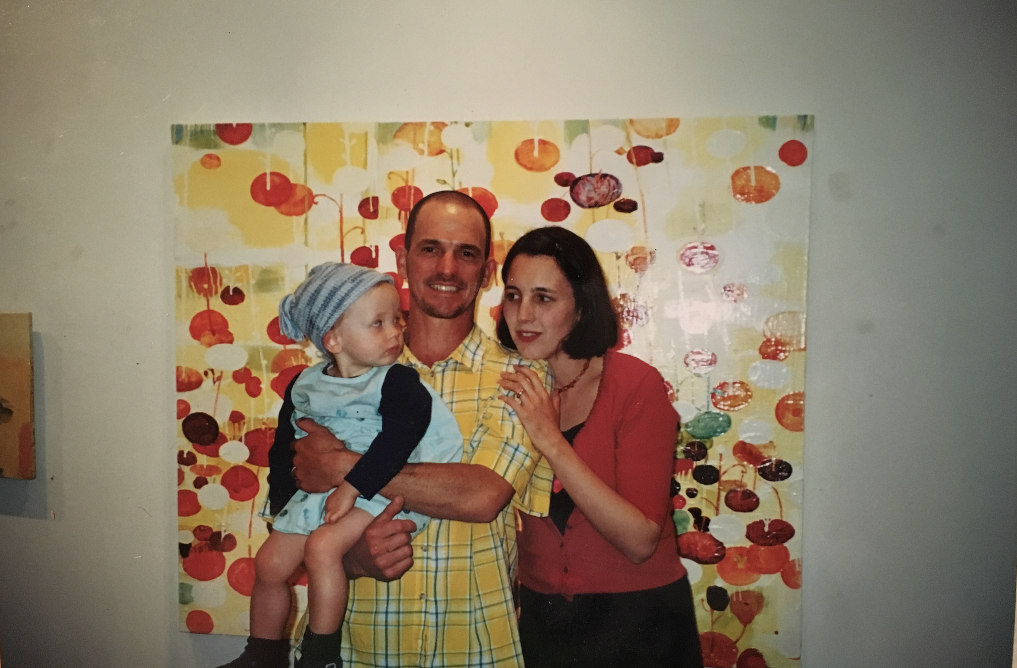 Ken holding their young daughter alongside Ellen in the 1990s, standing in front of one of Ken’s colorful oil paintings from his time as an artist before founding MAK.