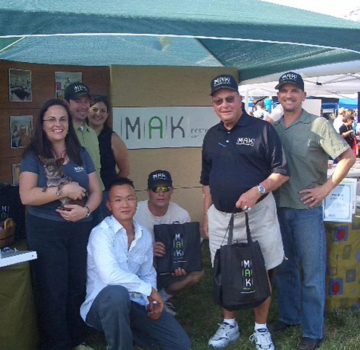 Early MAK Design + Build team posing at a community event booth with MAK signage, showcasing the company’s grassroots beginnings and connection to local clients.