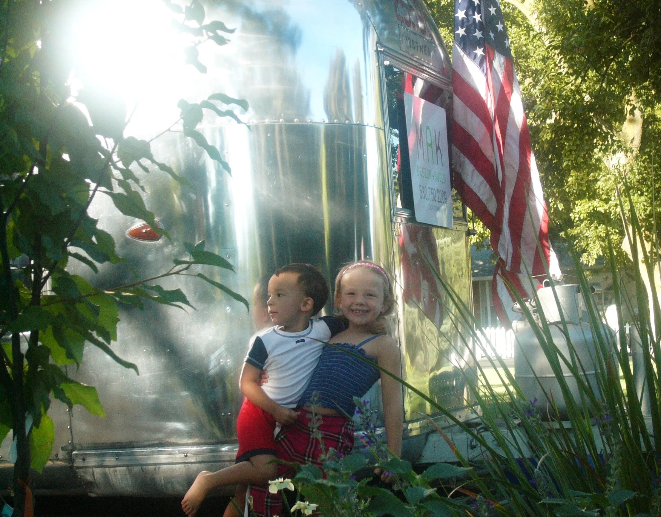 Ken and Ellen’s young children smiling in front of MAK’s original trailer office, a small silver Airstream that marked the early days of MAK Design + Build.”