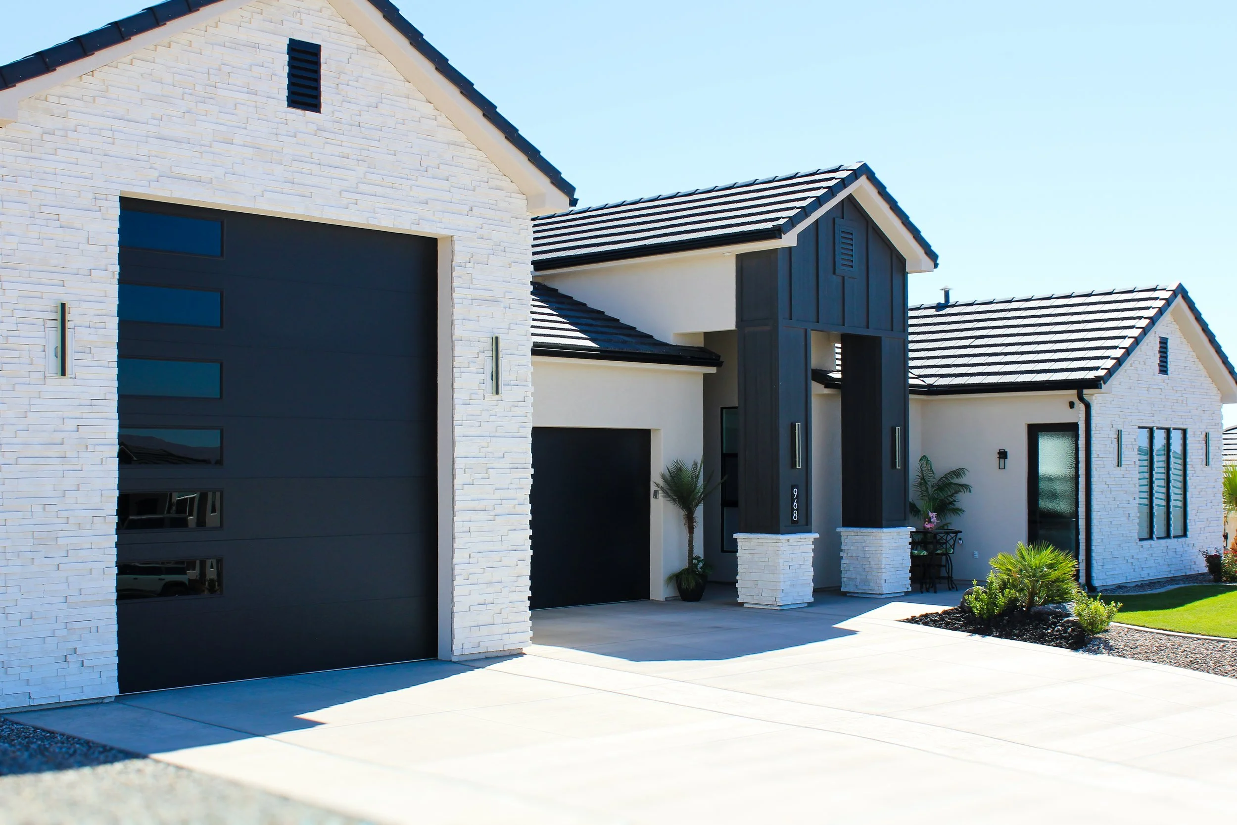 Modern house with white brick exterior, black garage door, and front porch, landscaped yard with small palms and green grass.