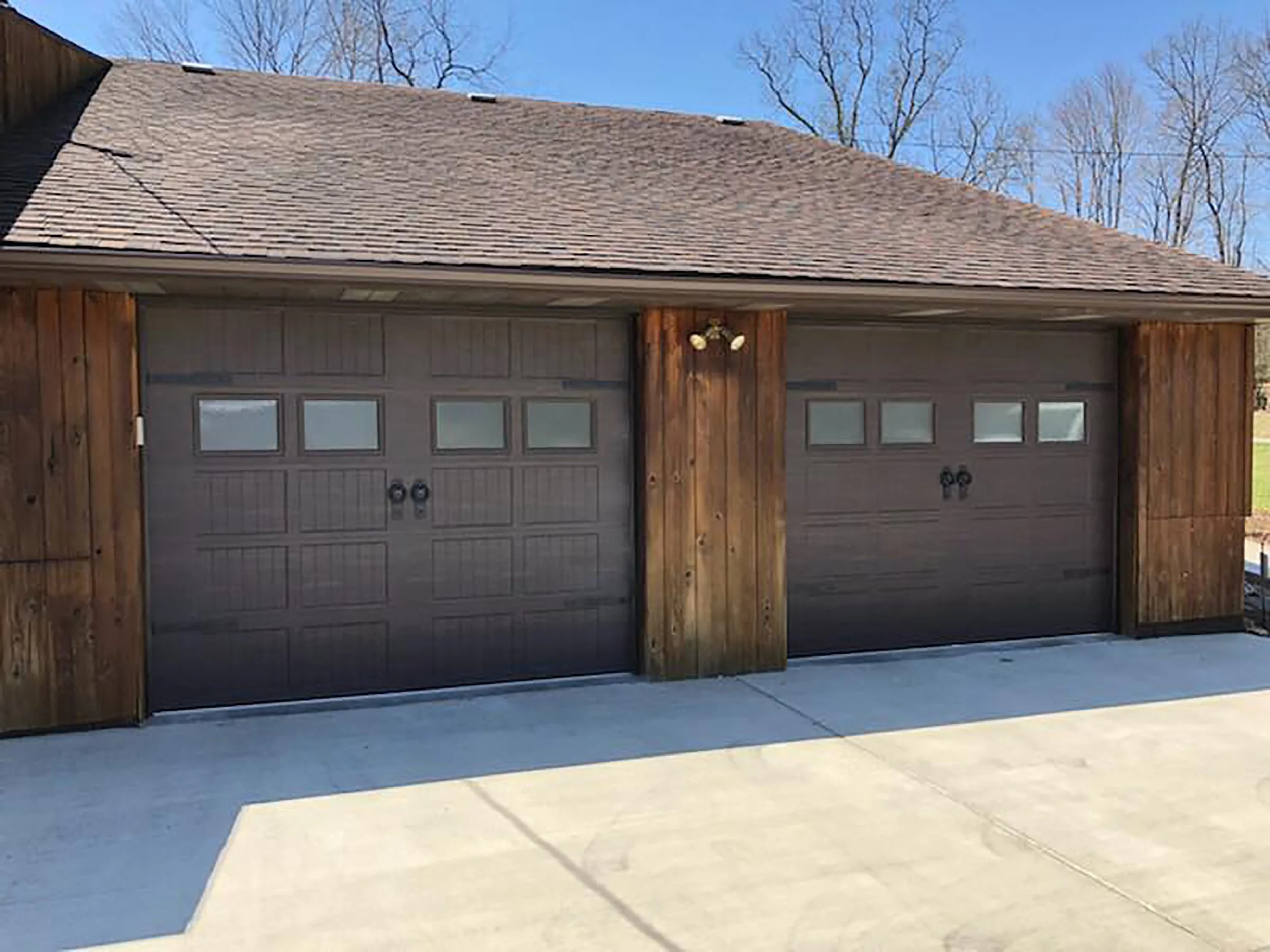 A detached garage with two brown doors, each with four small windows and black handles, surrounded by wooden siding and a sloped shingle roof, under a blue sky with leafless trees in the background.
