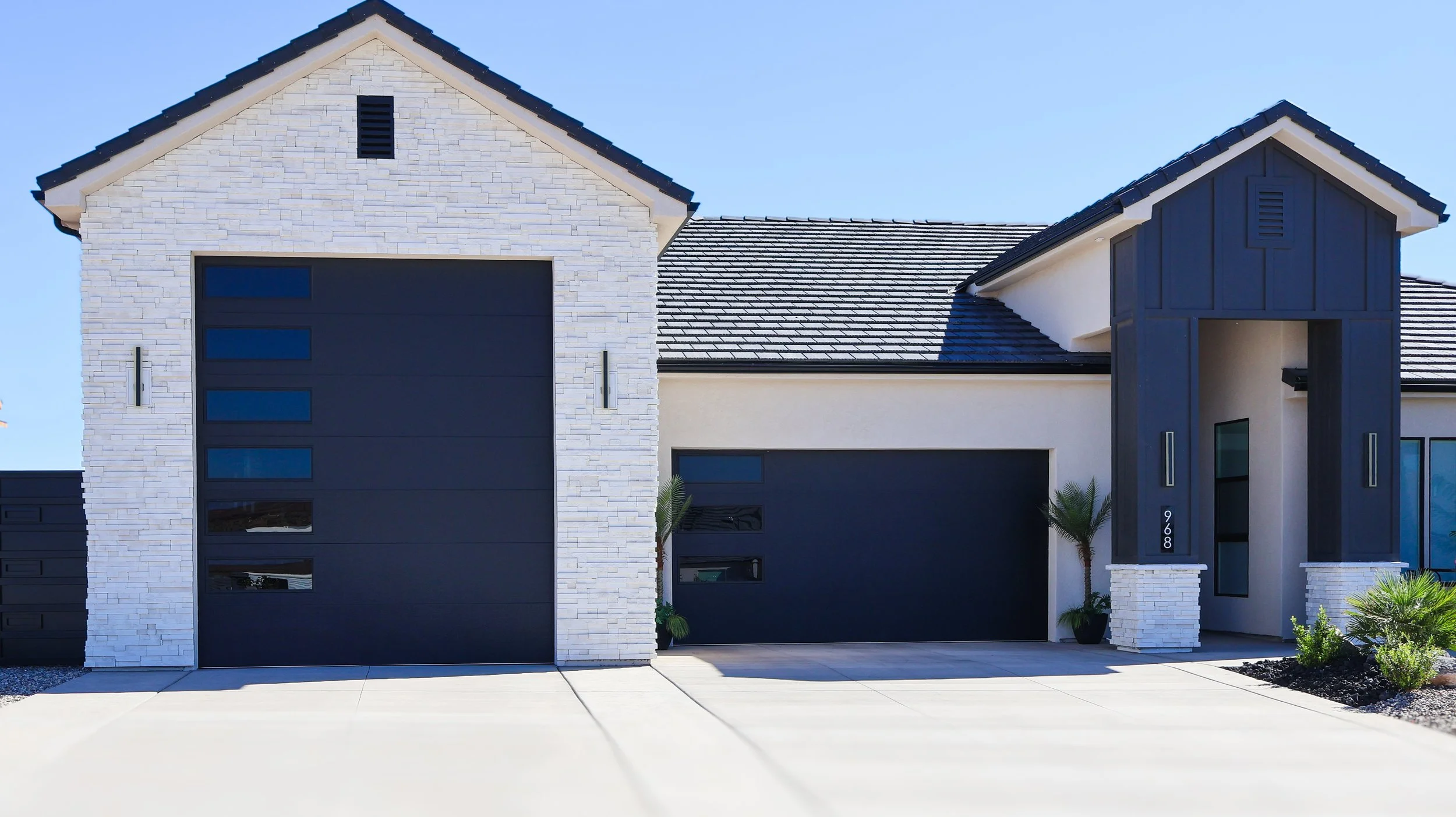 Modern house with white brick and dark gray accents, two large black garage doors, and small plants near the entrance.