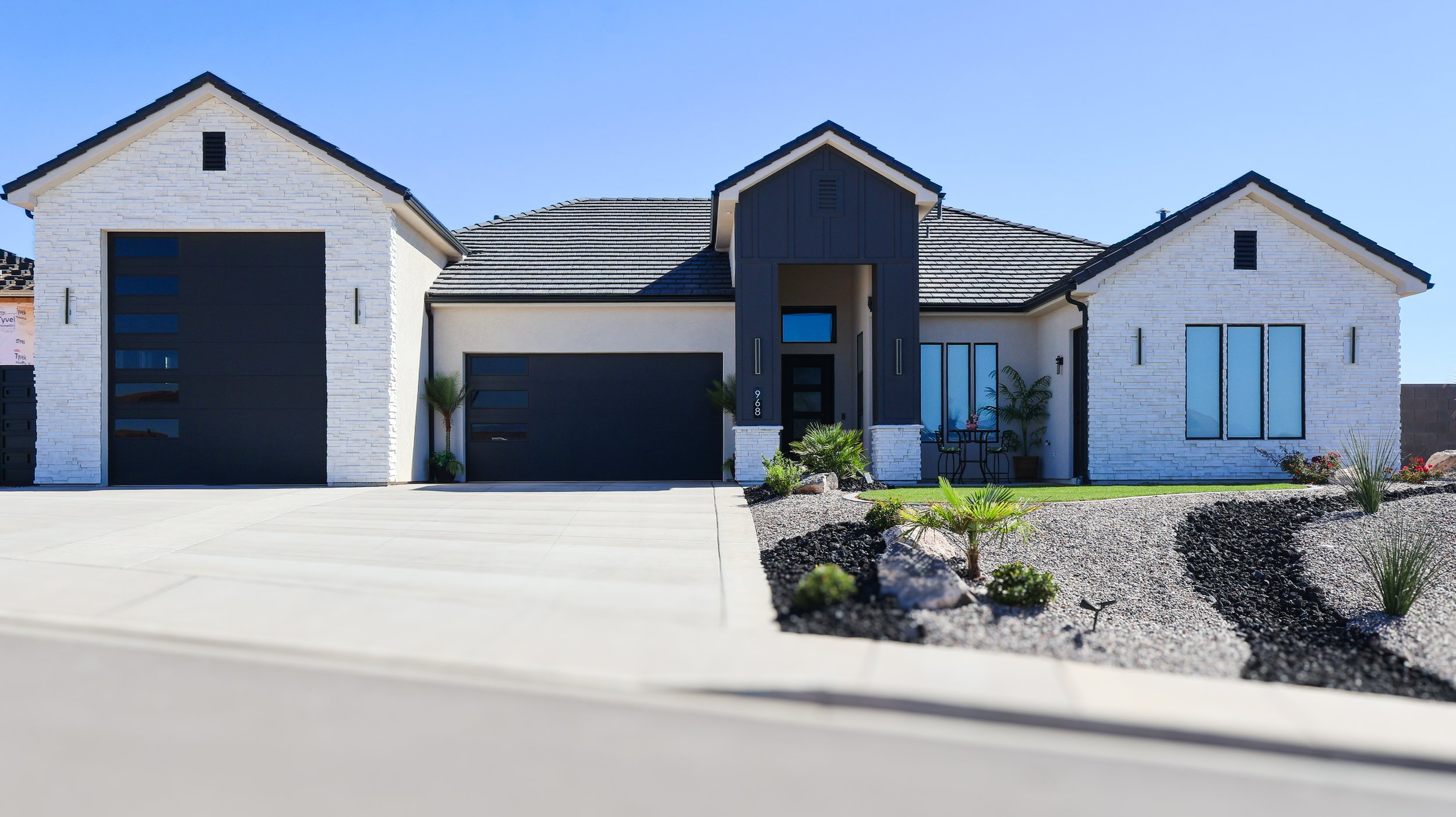 Front view of a modern house with a landscaped yard, white brick exterior, black garage doors, and a small sitting area outside.