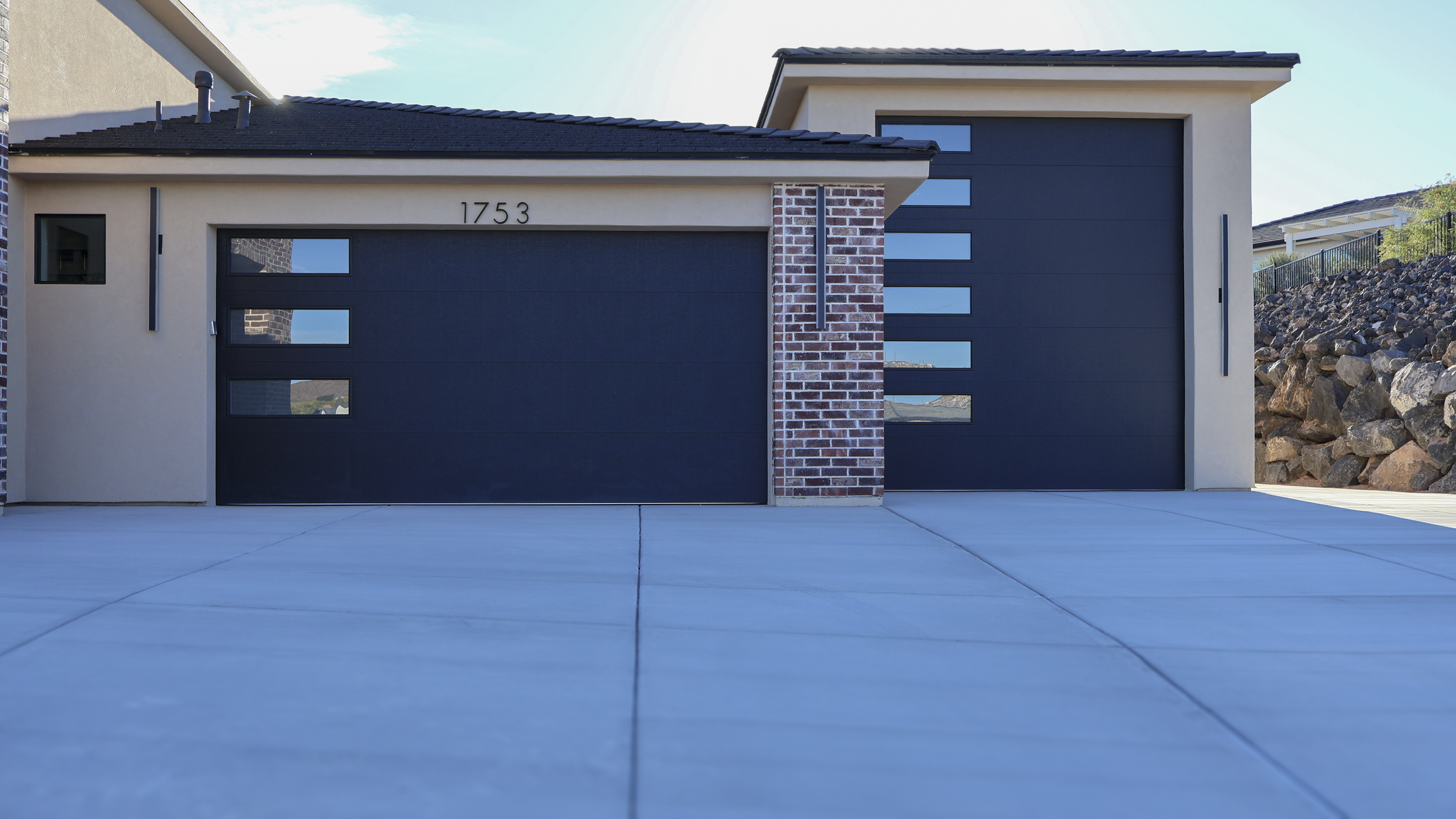 Modern house with two black garage doors, a beige exterior, and a brick pillar between the doors, with the house number 1753 above the left door. The driveway is concrete, and there is a rock hillside to the right.