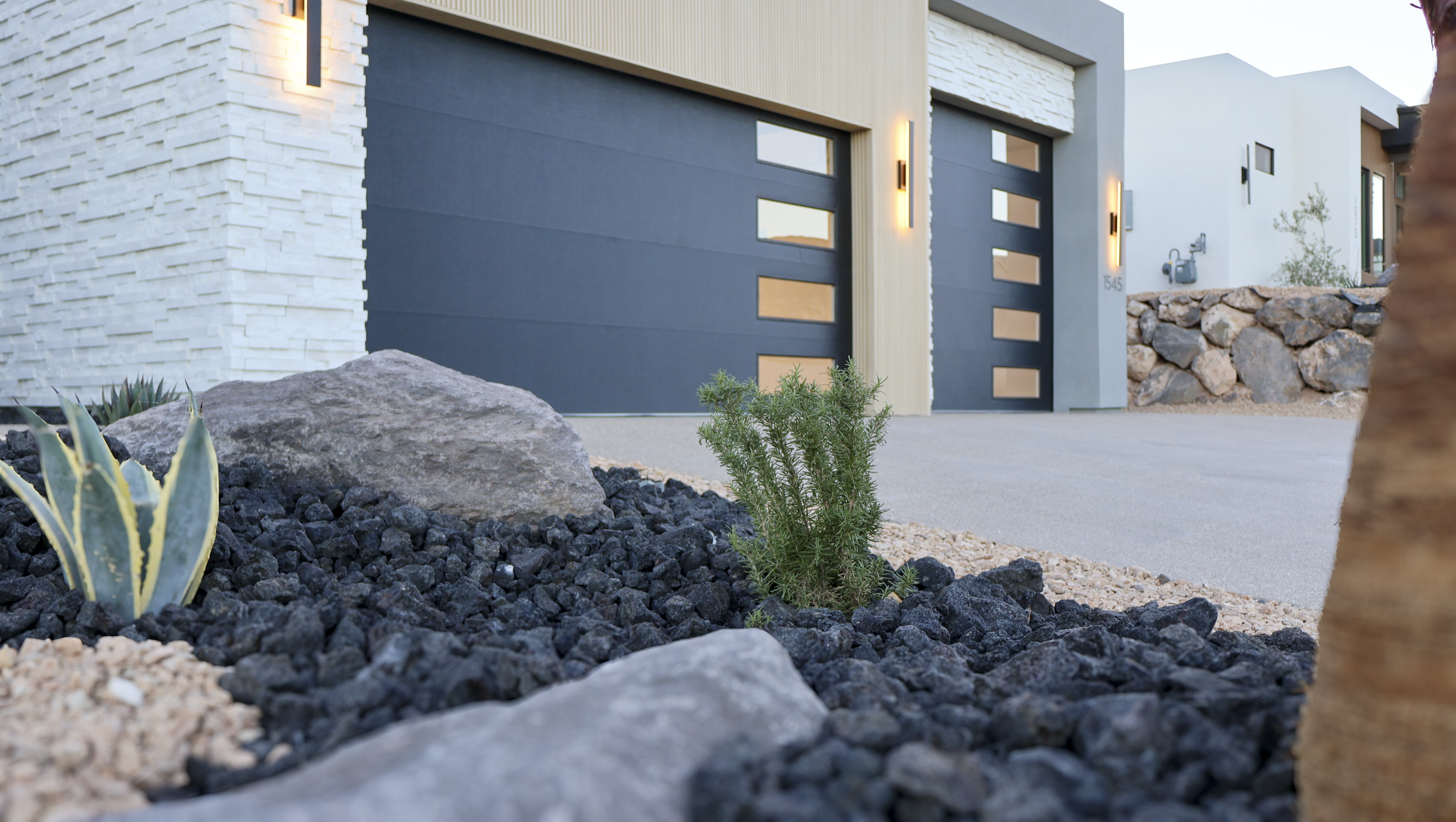 Modern house with dark gray garage door, white brick accents, and desert landscaping including rocks, succulents, and small shrubs.