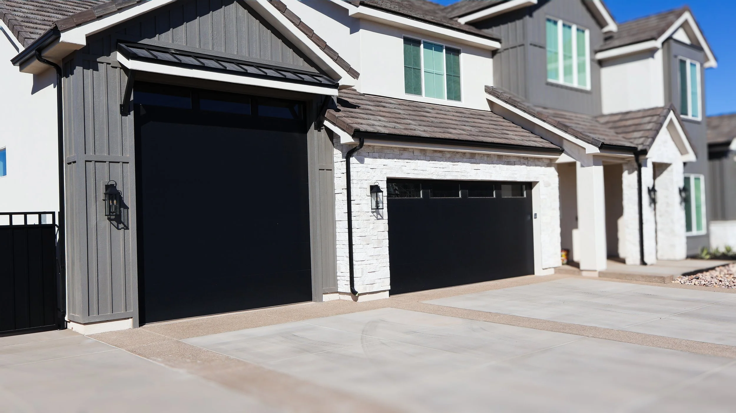 Exterior view of a modern house with two black garage doors, gray and white siding, stone accents, and two wall-mounted lanterns. The driveway is concrete.