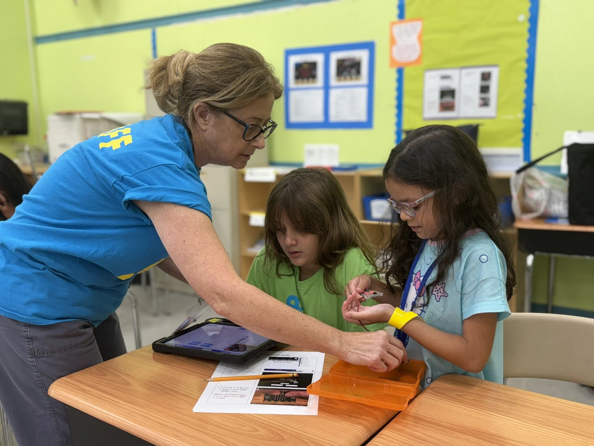 A teacher or adult assisting two young girls with an activity at a classroom table, surrounded by educational materials and a lime green classroom wall.