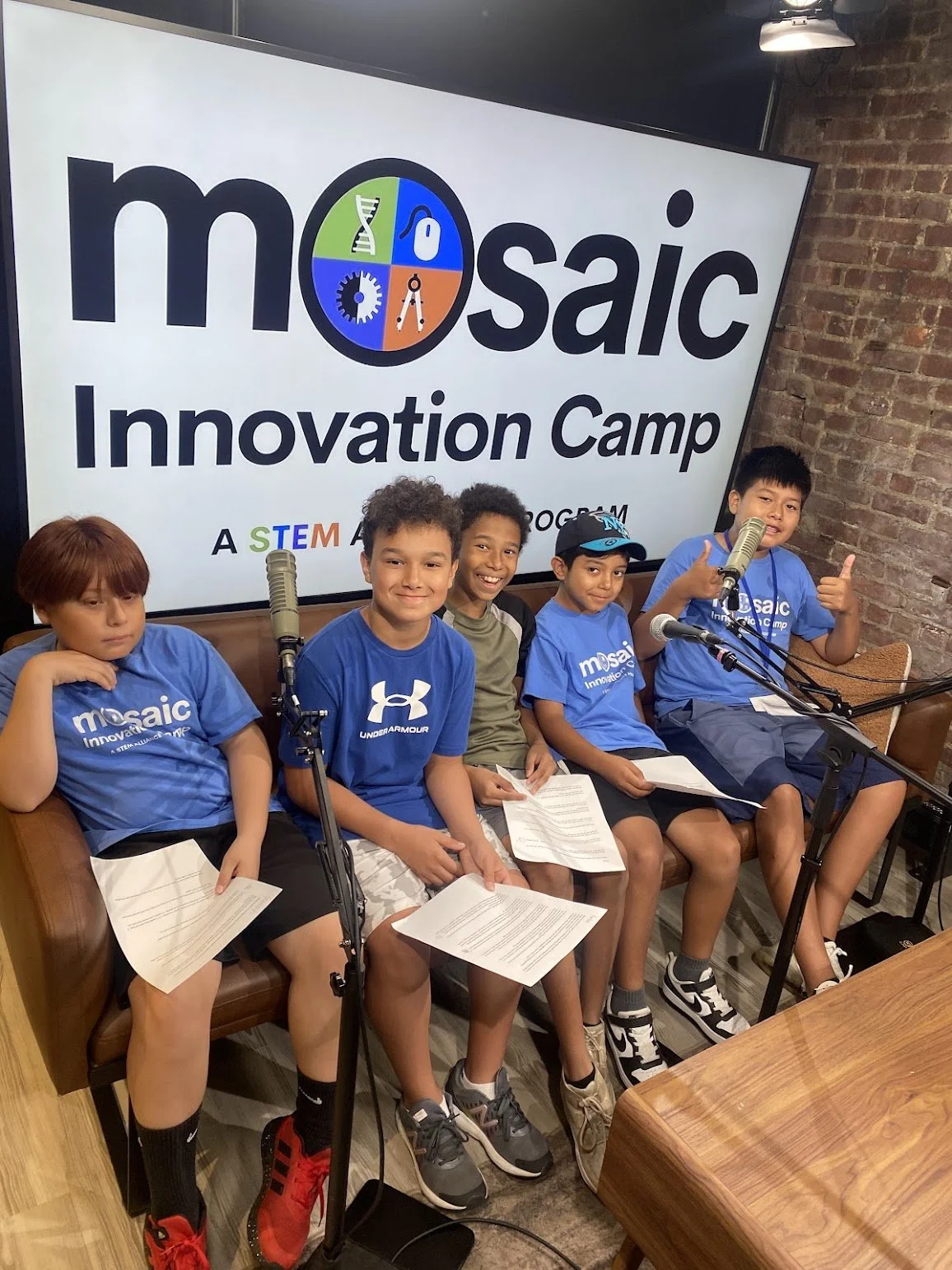 Five young boys in a recording studio with microphones sitting in front of a MOSAIC Innovation Camp banner