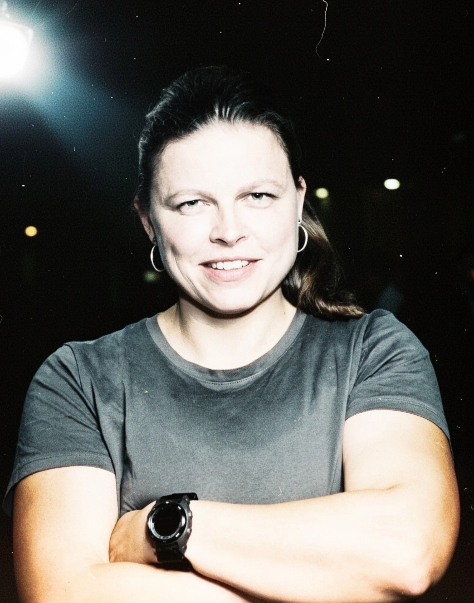Frau mit schwarzen Haaren, grauem T-Shirt, trägt eine schwarze Uhr und hat ihre Arme verschränkt, im Hintergrund ist Dunkel mit einigen Lichtern.