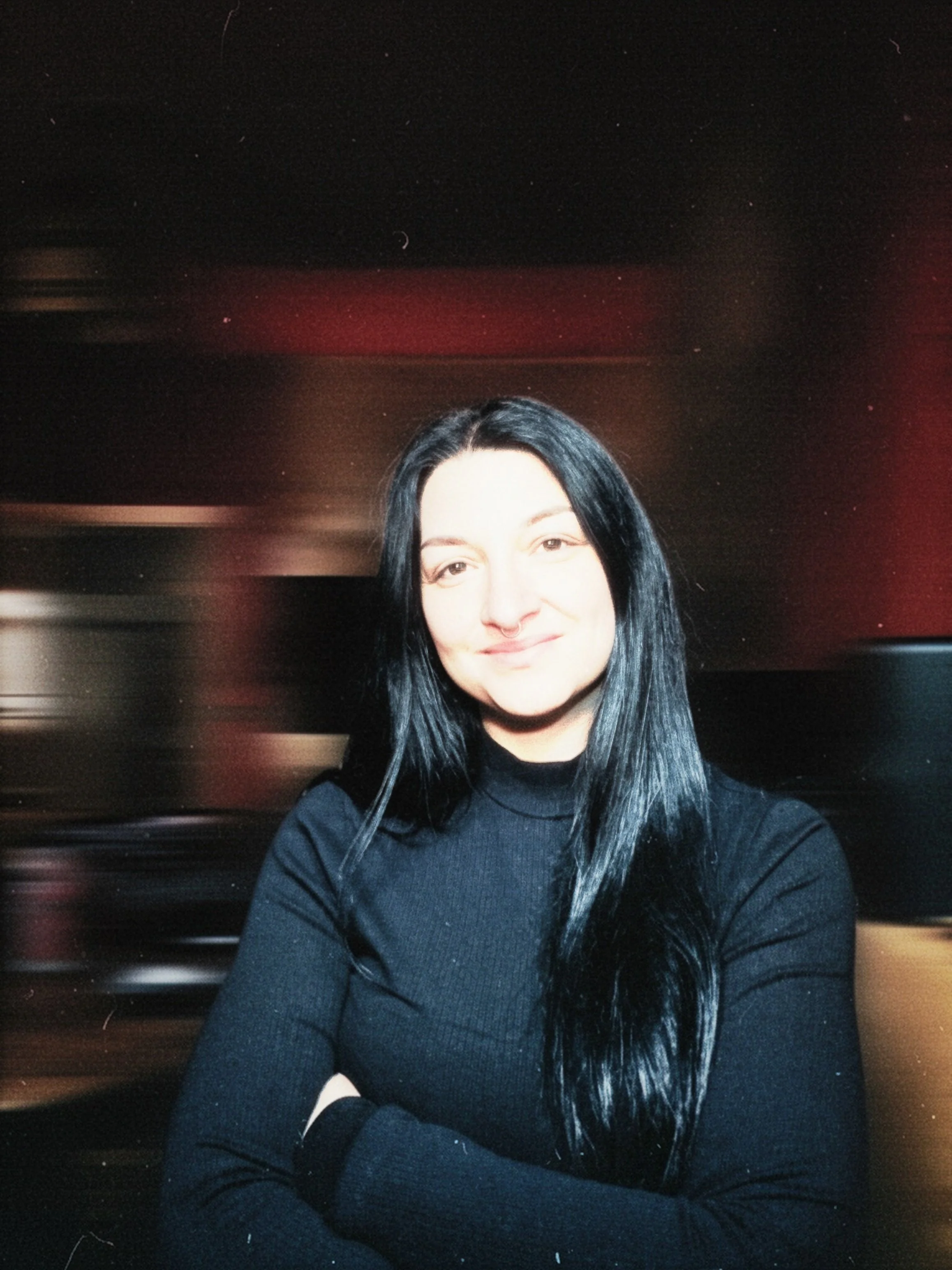 Junge Frau mit langen schwarzen Haaren und schwarzem Pullover steht vor einem verschwommenen Hintergrund mit dunklen und roten Farben, lächelnd mit verschränkten Armen.