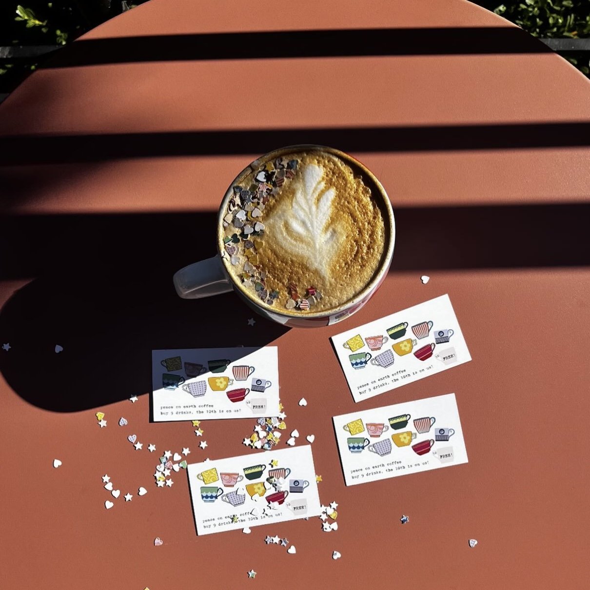 A cup of coffee with latte art and colorful sprinkles, placed on a pink surface. Surrounding the cup are three small printed coupons with il