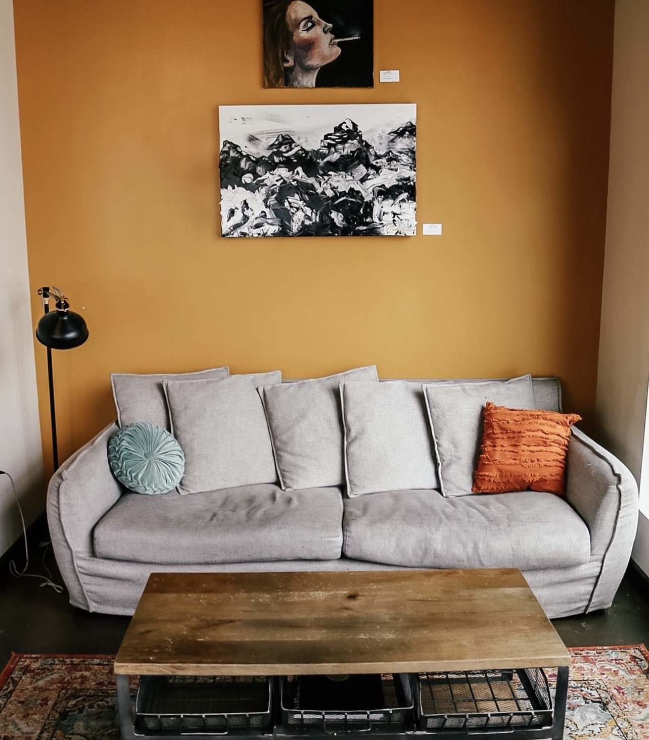 Living room with a beige sofa, orange and teal pillows, a wooden coffee table, a black floor lamp, and orange/white wall art.