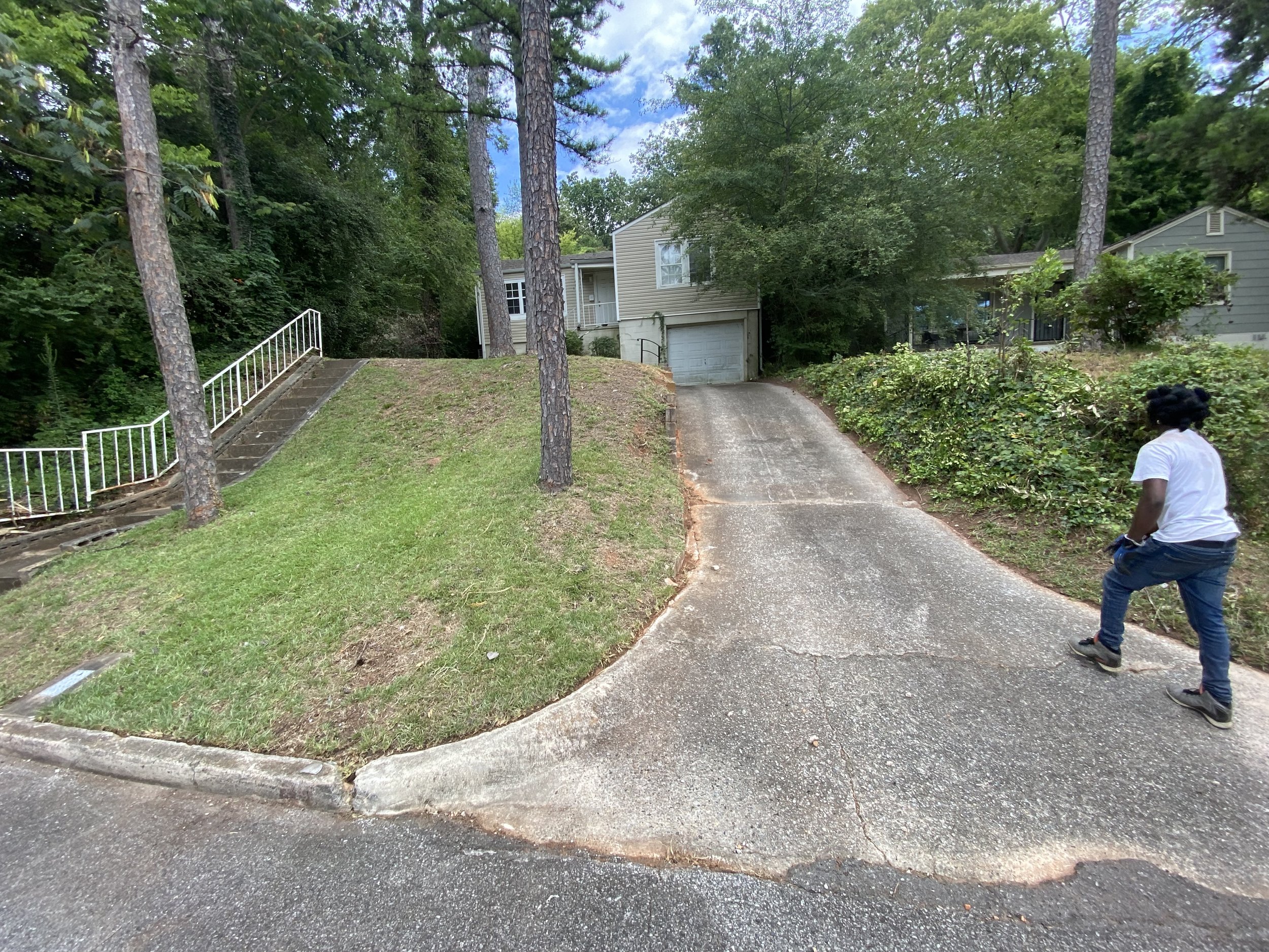A man in a white shirt and blue jeans walking on a paved driveway leading to a house with a garage, surrounded by trees and greenery.