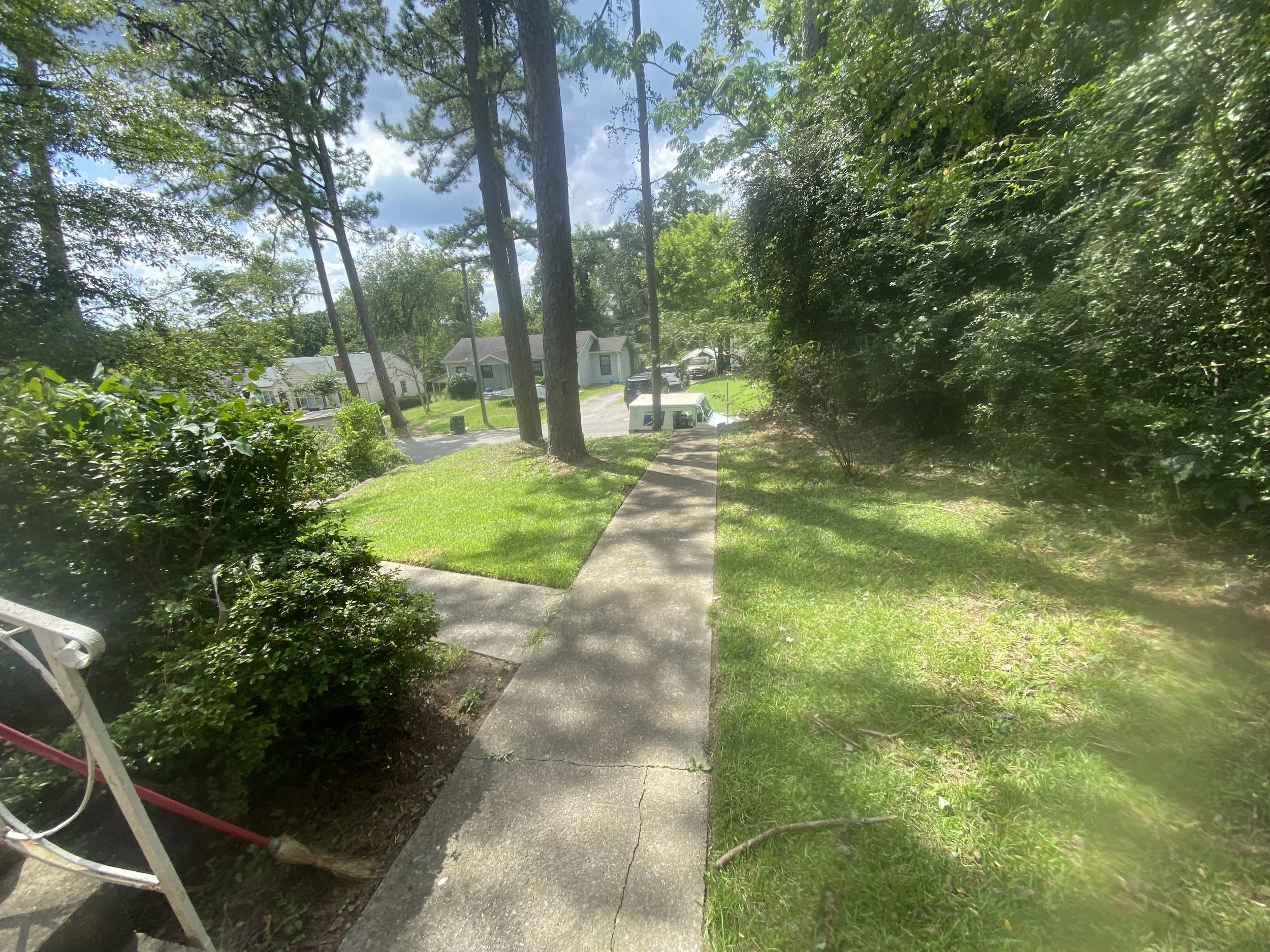 Sidewalk leading to a residential area with trees and houses in the background, sunny day with partly cloudy sky.