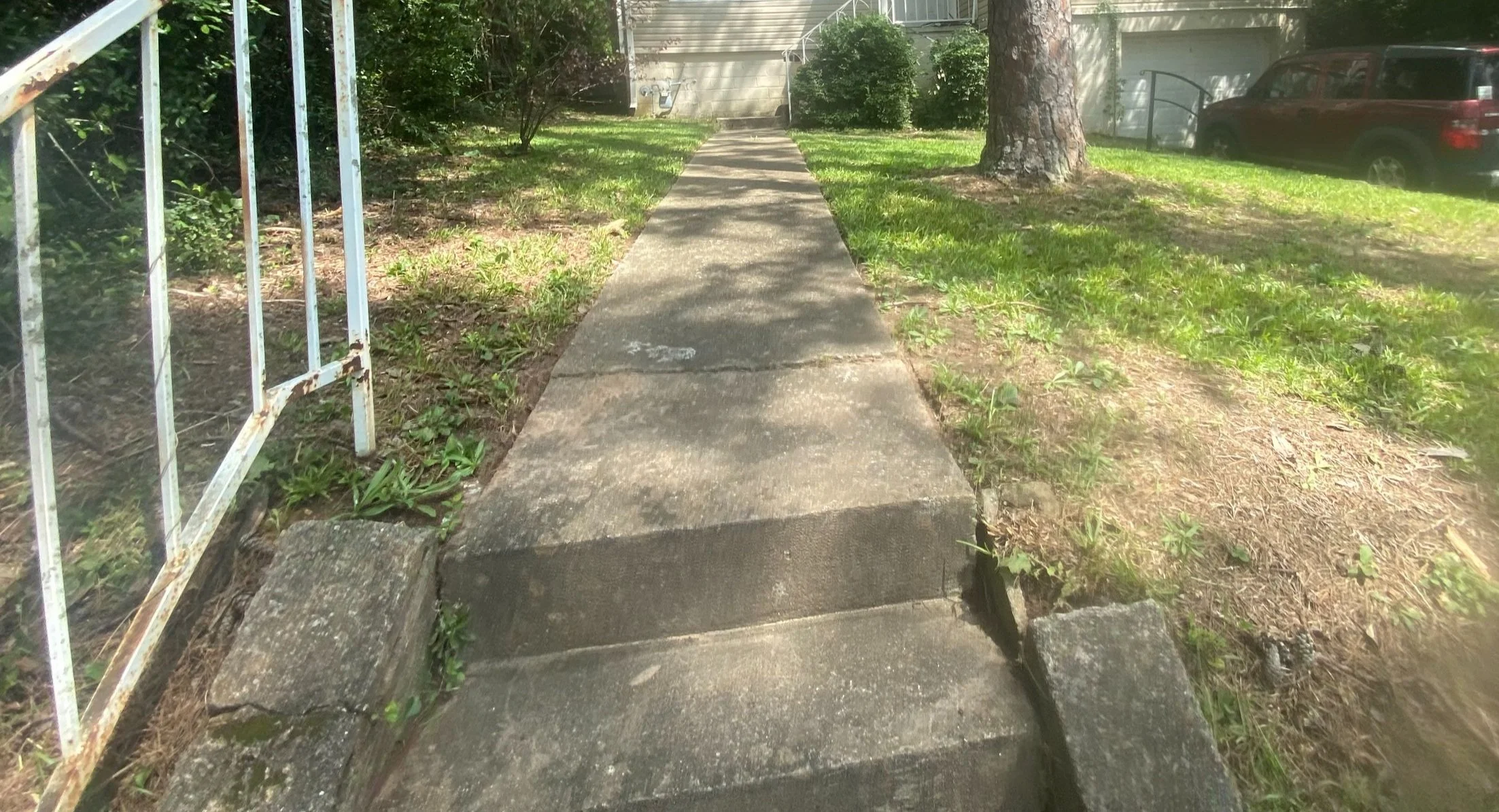 Concrete sidewalk with cracks and patches, leading towards a house in the background, flanked by grassy areas, mature trees, and a parked red vehicle.