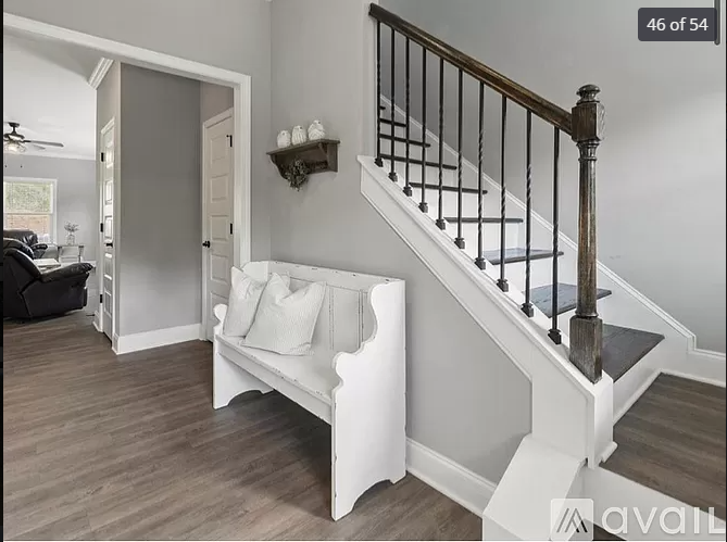 Interior view of a home's entryway with a staircase, a white wooden bench with pillows, a small wall shelf with decorative items, and a doorway leading to a living room with a black sofa and large windows.