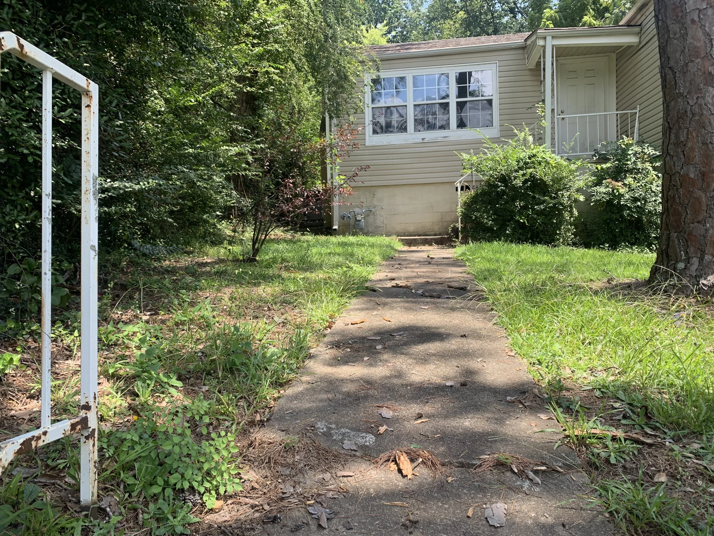 A sidewalk leading to a house with a small balcony on the right, surrounded by green bushes and trees, with a metal front gate on the left.