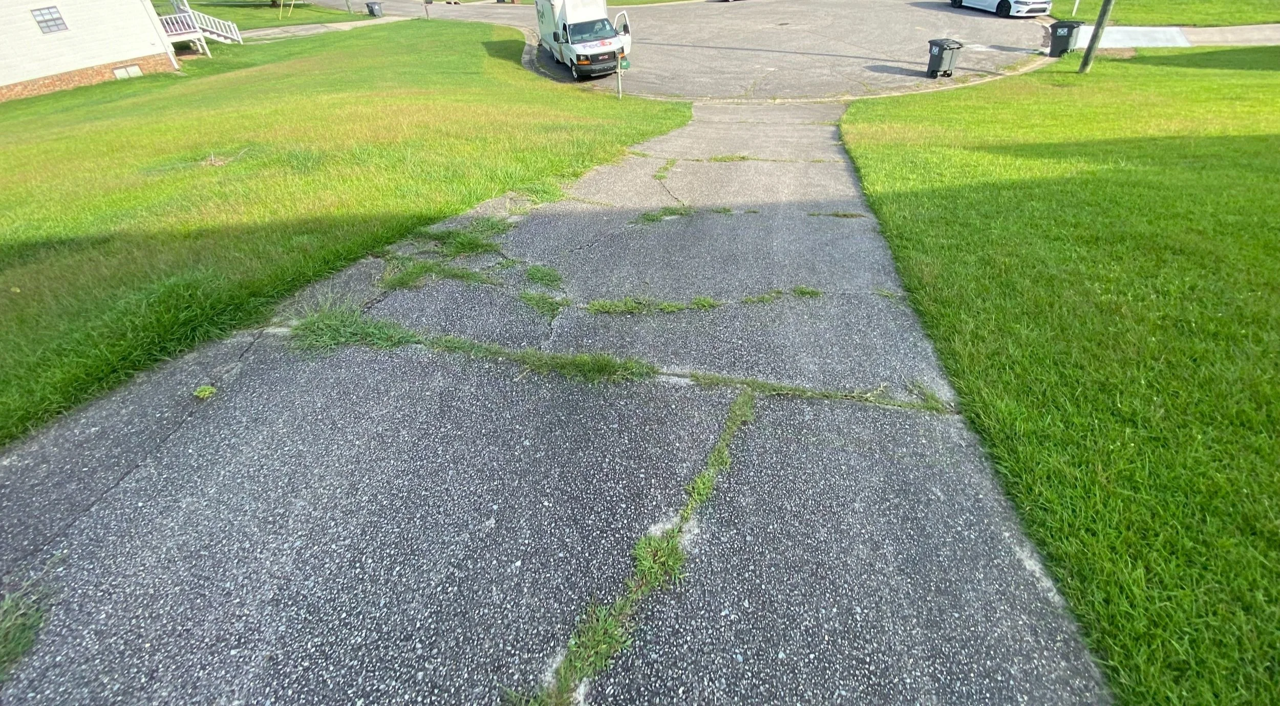 Cracked sidewalk with grass growing in the cracks, green lawns on sides, and a driveway with parked vehicles in the background.