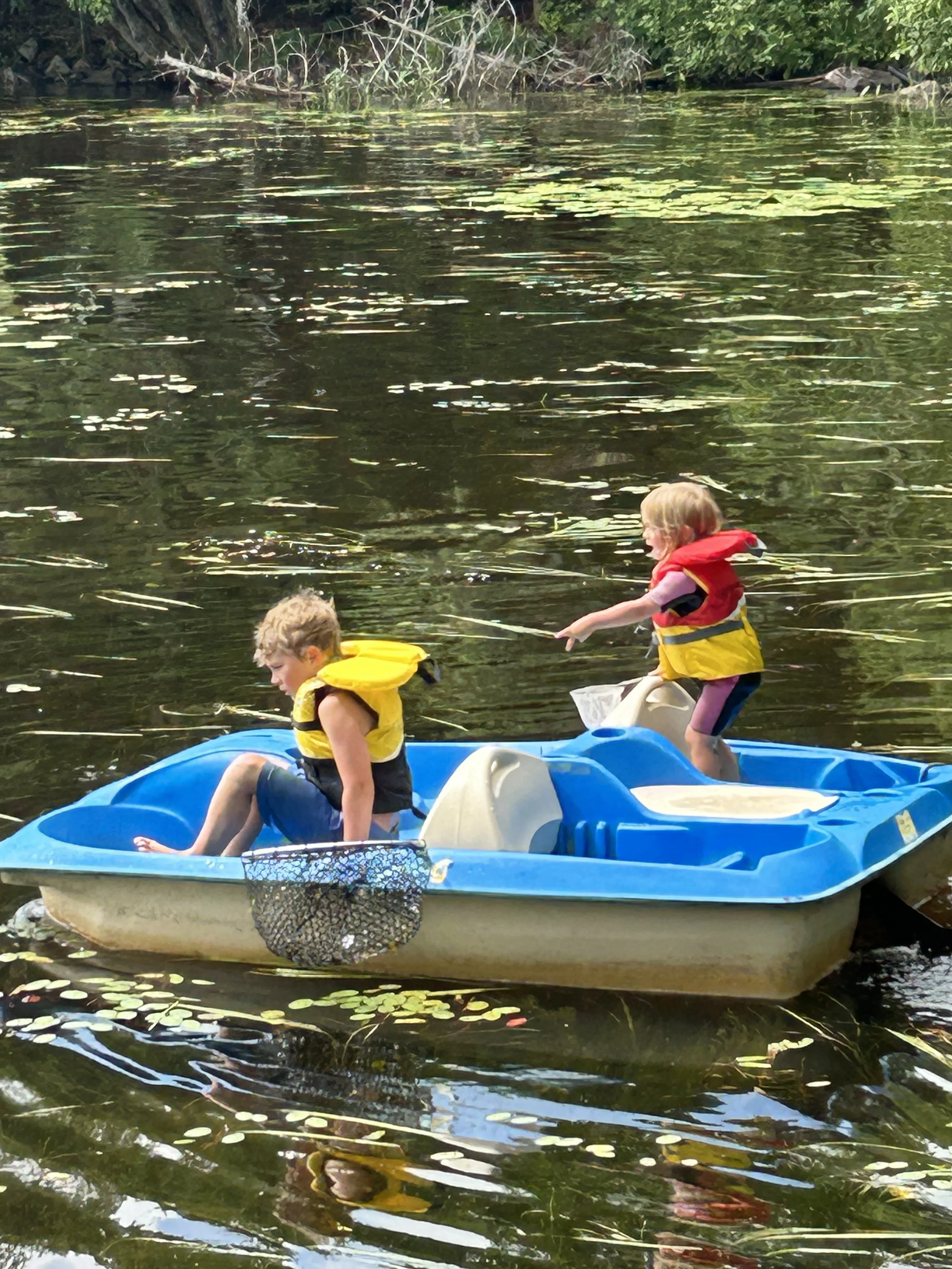 Two children in a blue paddle boat on a lake, both wearing life jackets, with one sitting and the other standing and reaching out with a pole.